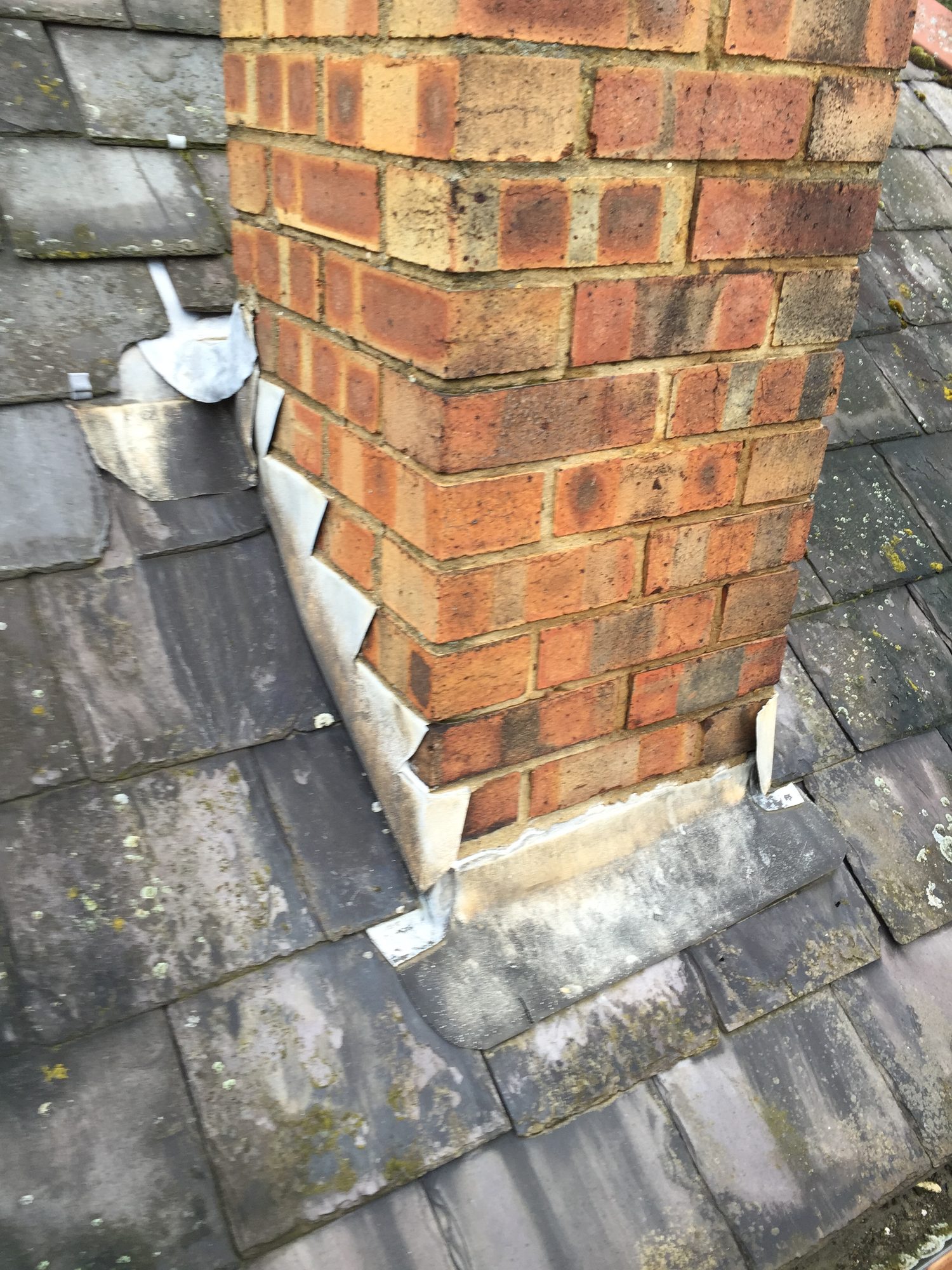 Brick chimney surrounded by dark slate roof tiles, with sections of metal flashing visible around the base of the chimney