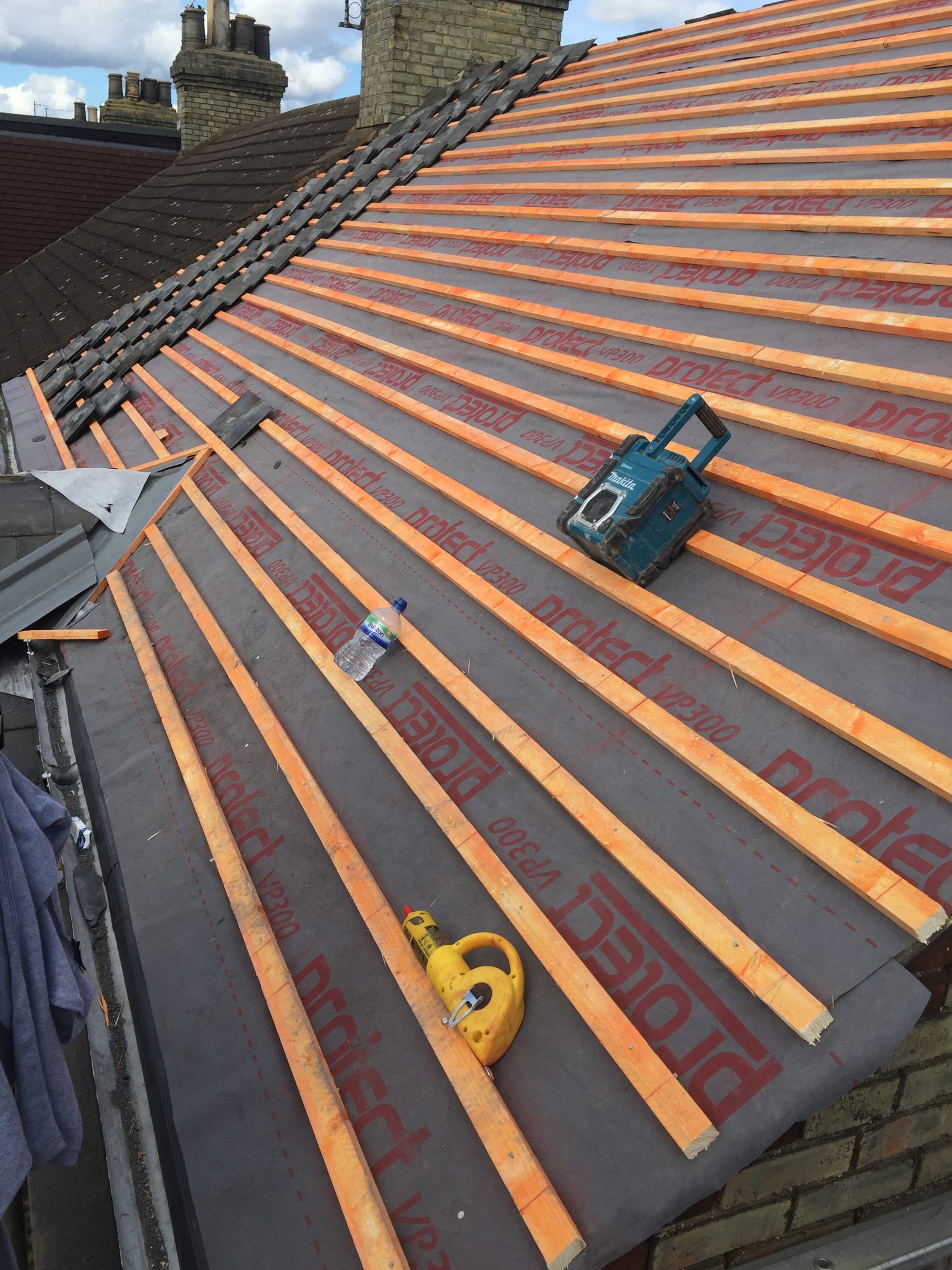 Sloping roof showing grey roofing underlay and timber battens installed across the roof, with roofing tools and materials visible on the surface