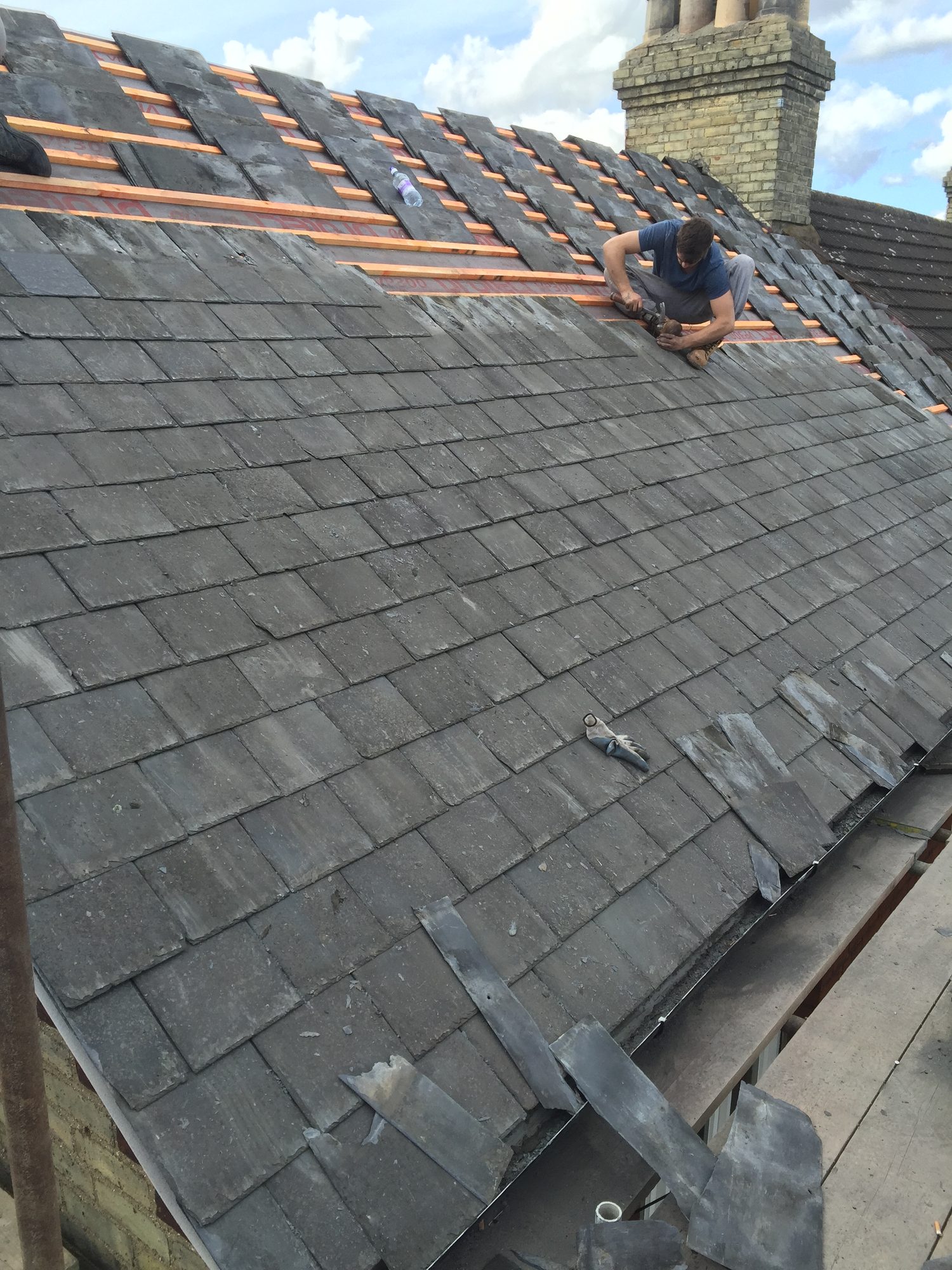 Sloping roof with rows of dark slate tiles installed across the lower section while a worker installs further tiles on the upper section, with roofing materials visible nearby