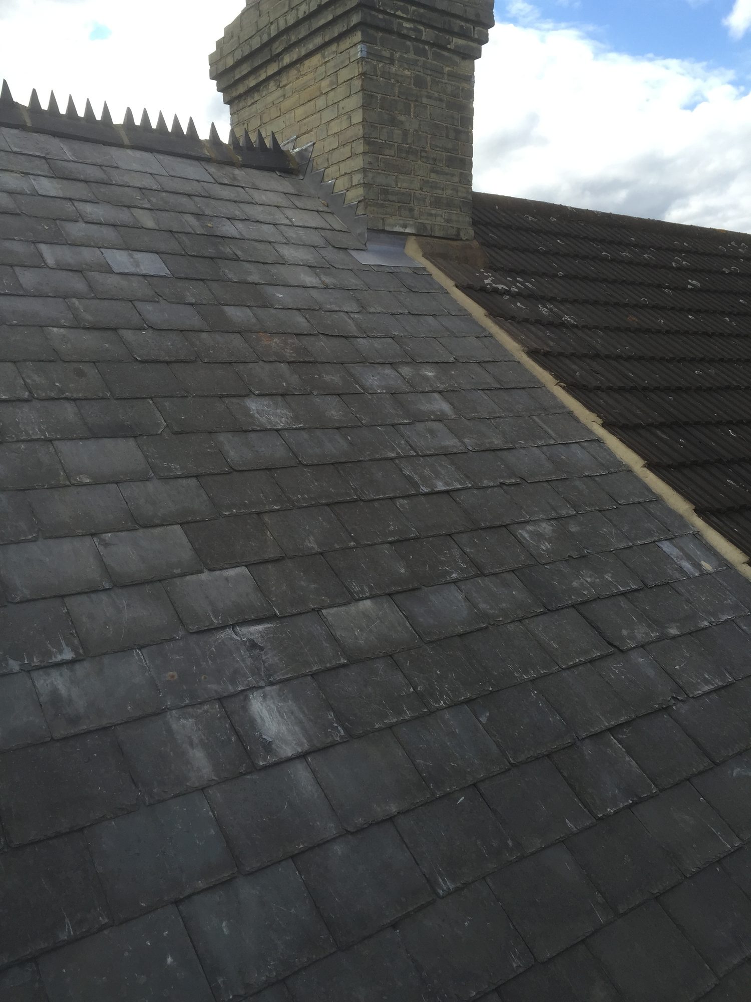 Completed sloping roof covered with rows of dark slate tiles, with the renewed roof meeting a brick chimney and adjoining tiled roof section