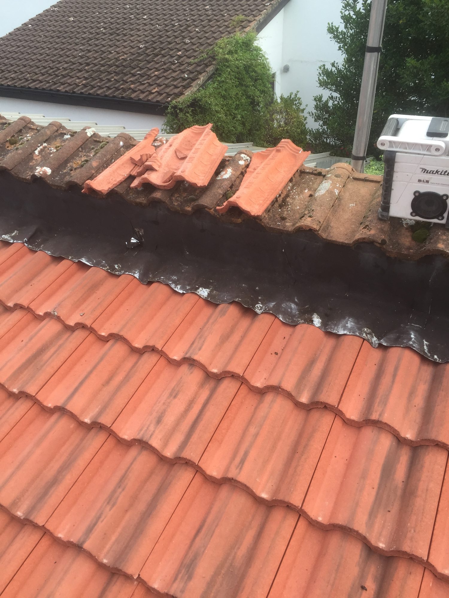 Close view of a tiled roof junction with dark leadwork running along the edge and several loose tiles positioned near the ridge, with surrounding roof equipment visible