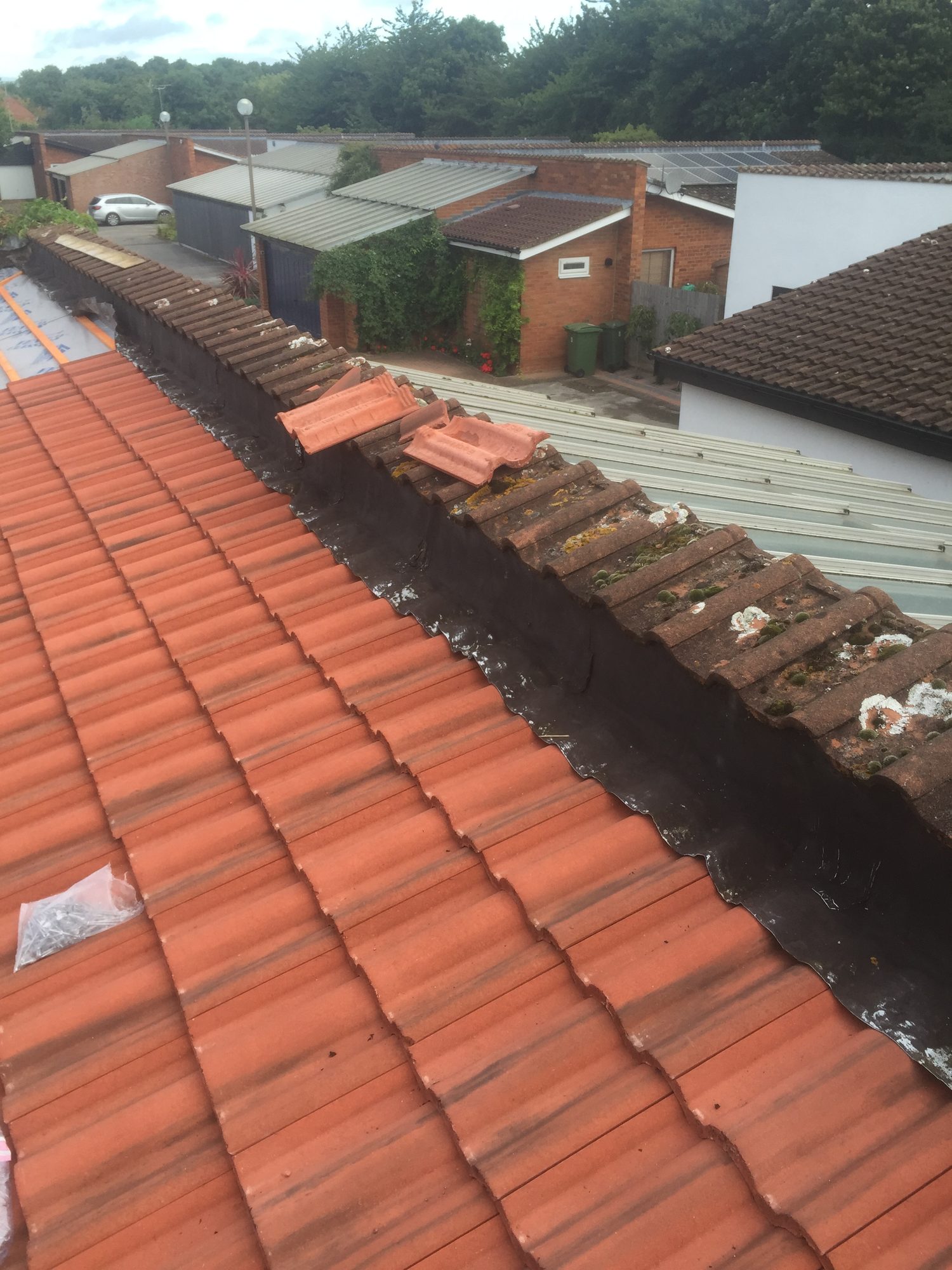 Wider view of a tiled roof showing the roof junction, dark leadwork and moss-covered tiles, with neighbouring buildings and trees visible in the background