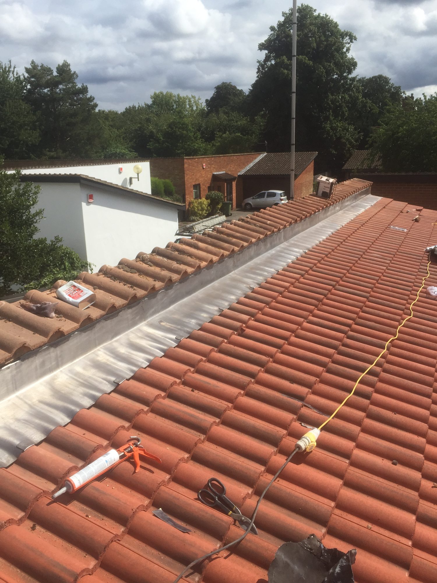 Wide view of a tiled roof showing newly installed leadwork along the junction with the adjoining raised roof section, with tools and materials visible on the tiles