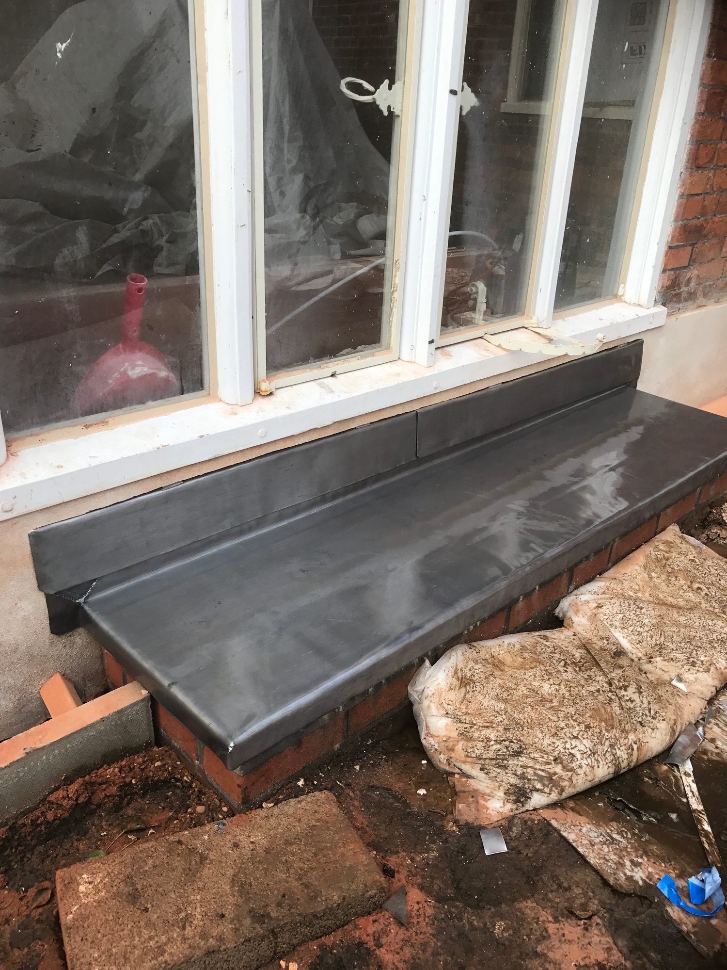 Finished dark grey leadwork covering a brick bay beneath white-framed windows, with formed edges, seams and detailing visible around the roof perimeter