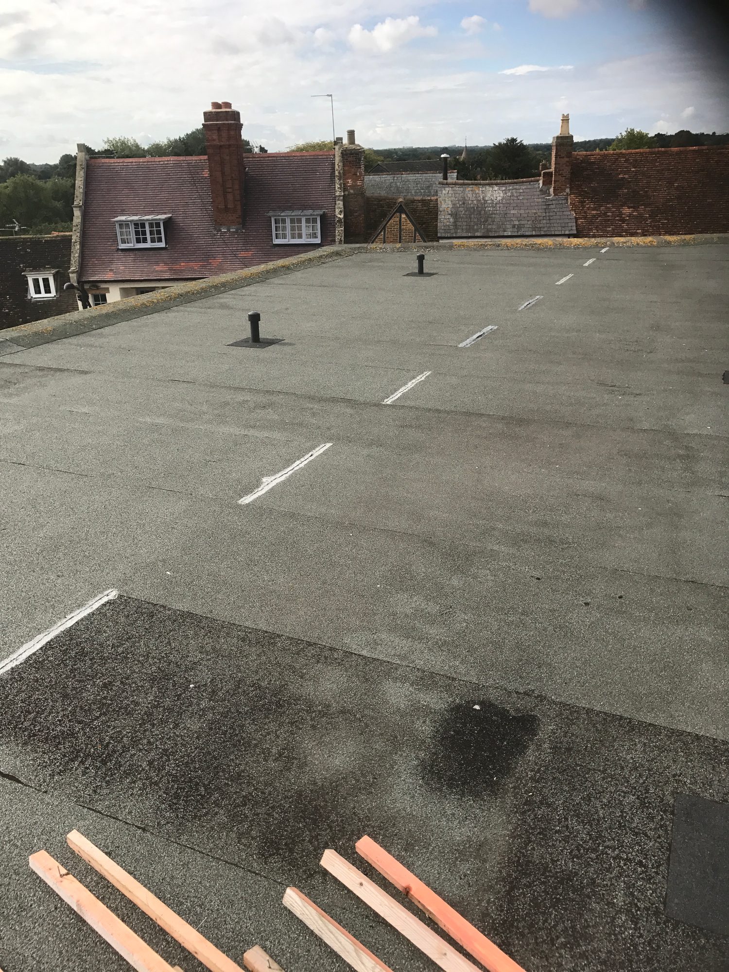 Existing flat roof covered with a grey surface, showing several roof outlets and marked seams, with timber battens visible in the foreground and neighbouring tiled roofs beyond