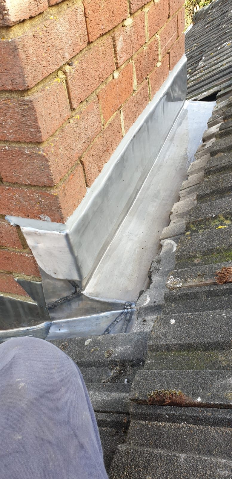 Close-up of lead flashing formed against a brick wall and adjoining roof covering, with folded and jointed sections visible at the roof junction