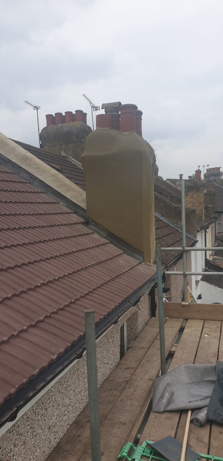 View of a roof chimney with lead flashing visible along its base and sides, with tiled roof coverings, scaffolding and neighbouring chimneys in the background