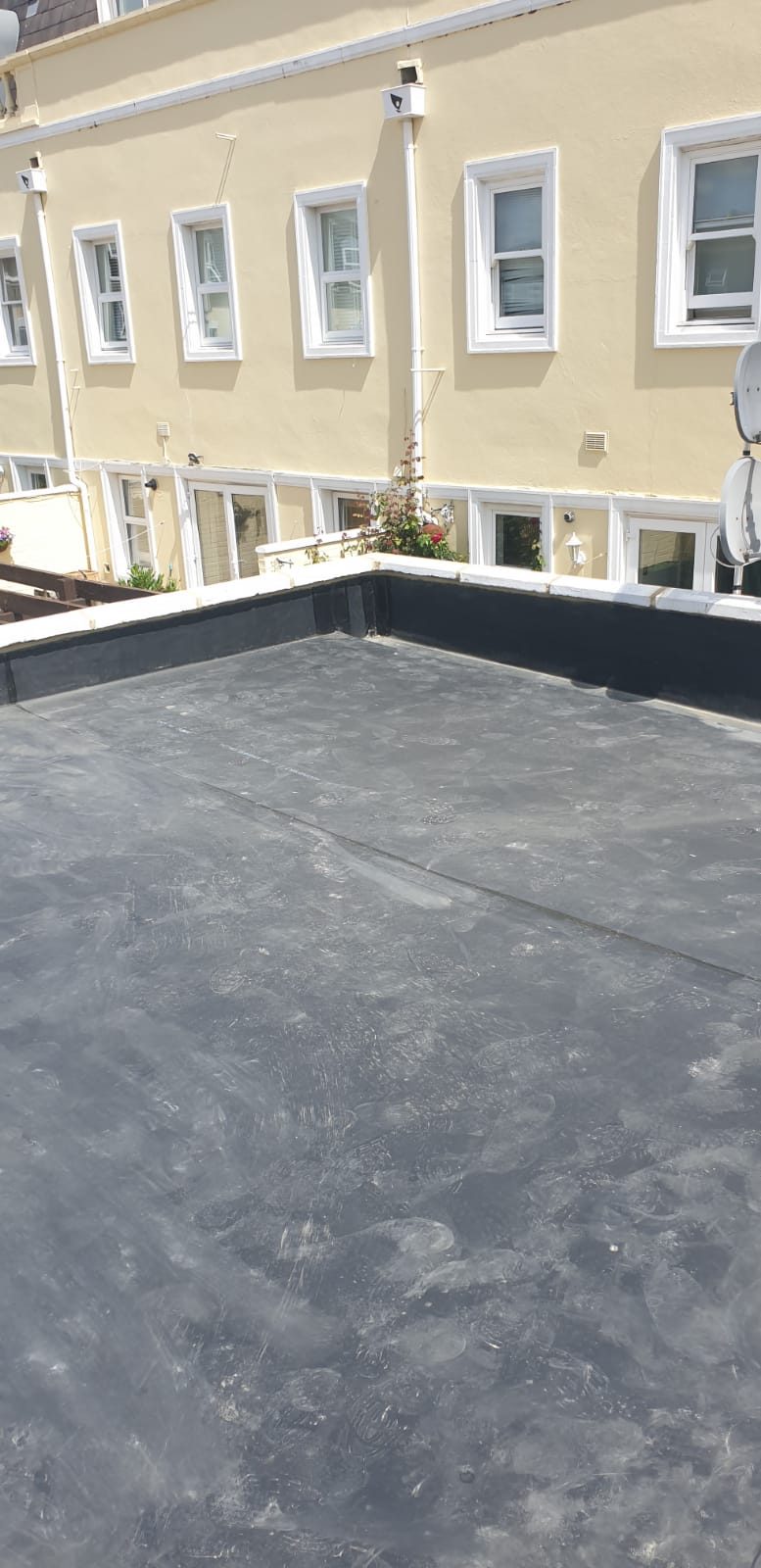 View across the finished flat roof showing the dark grey roof surface, perimeter upstands and light-coloured coping stones, with the adjoining residential buildings visible beyond
