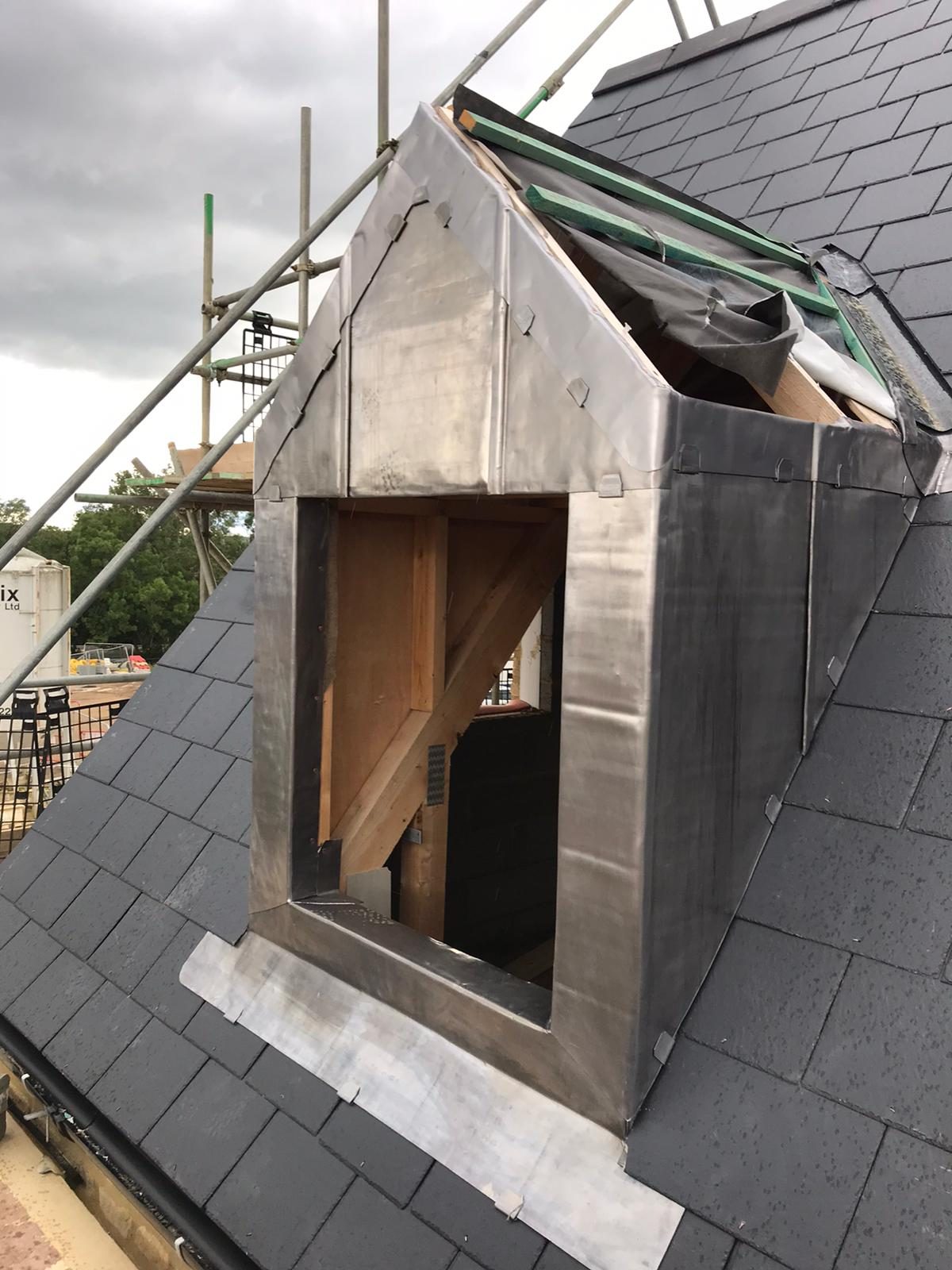 Close view of a dormer clad in lead with neatly formed seams and fixings, set into a dark slate roof with timber framing visible through the opening