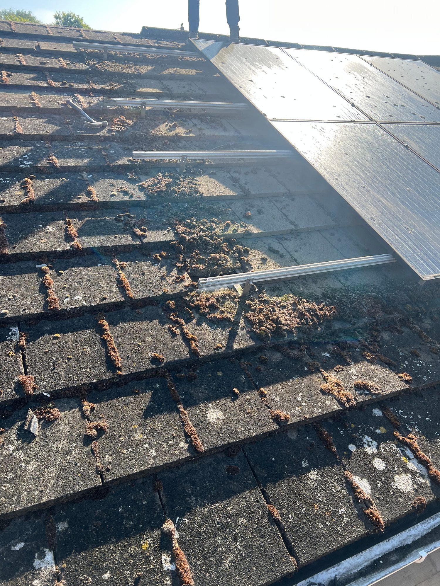 Close view of weathered roof tiles with solar panels and aluminium mounting rails installed, with a worker’s legs visible at the top of the roof