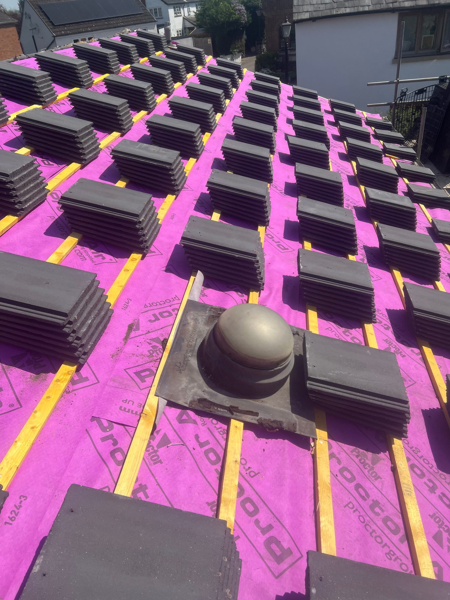 Roof covered with bright pink roofing membrane and timber battens, with stacks of dark roof tiles arranged across the surface and a roof vent visible