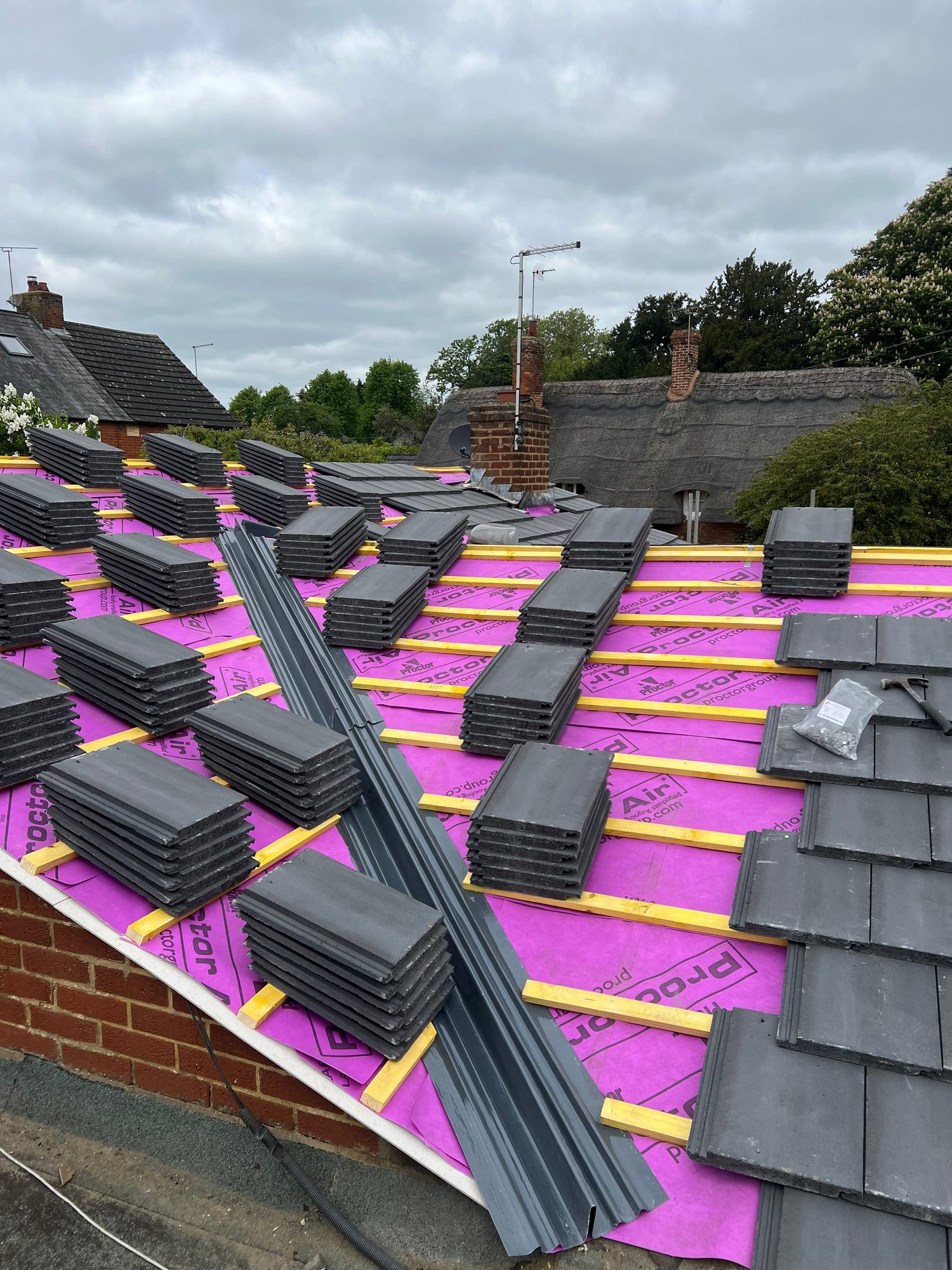 Wide view of a roof covered with pink roofing membrane and timber battens, with multiple stacks of dark roof tiles and lengths of roof trim positioned across the surface