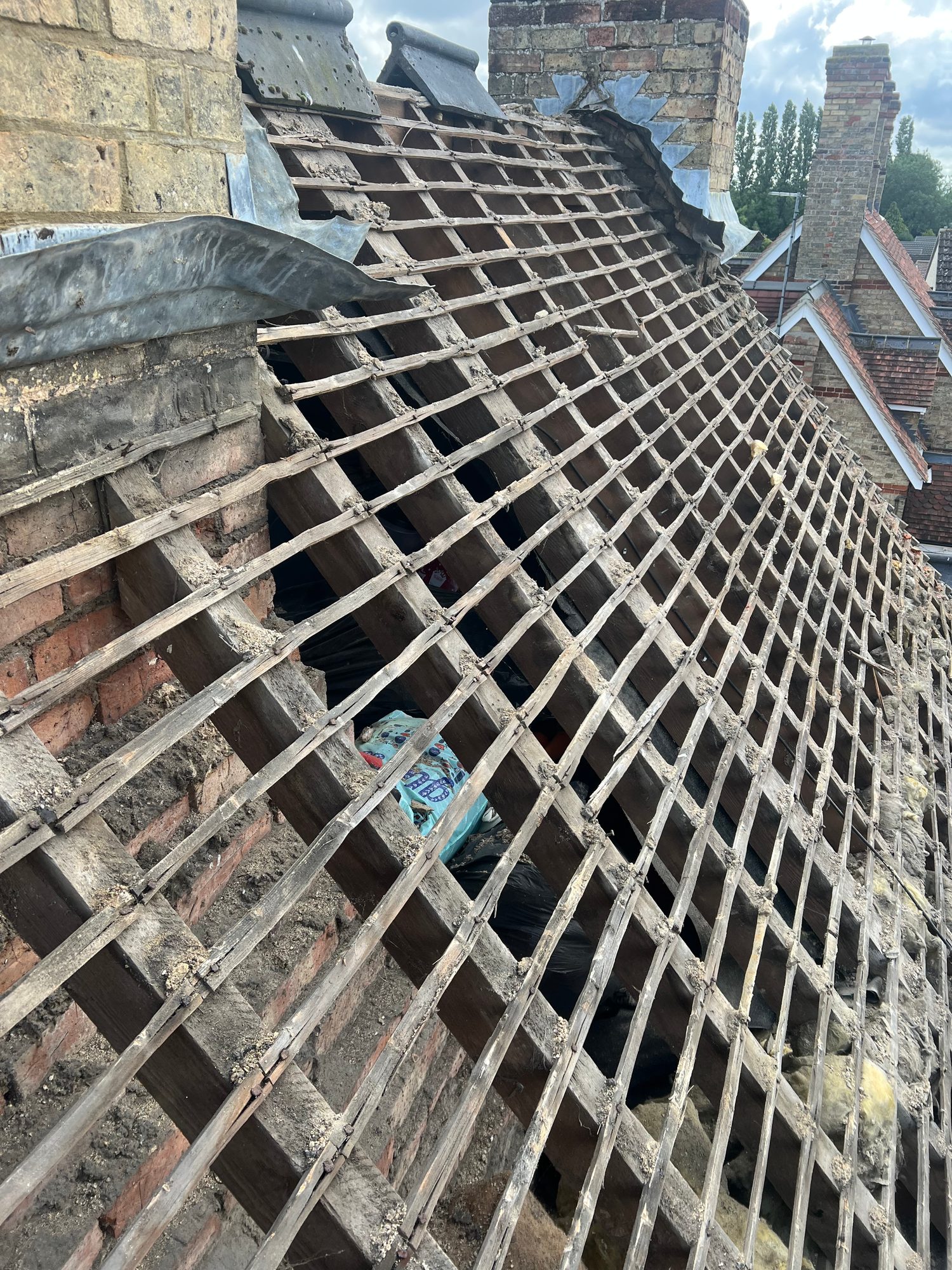 Close view of an exposed pitched roof showing timber battens, roof timbers and sections of damaged covering around a chimney