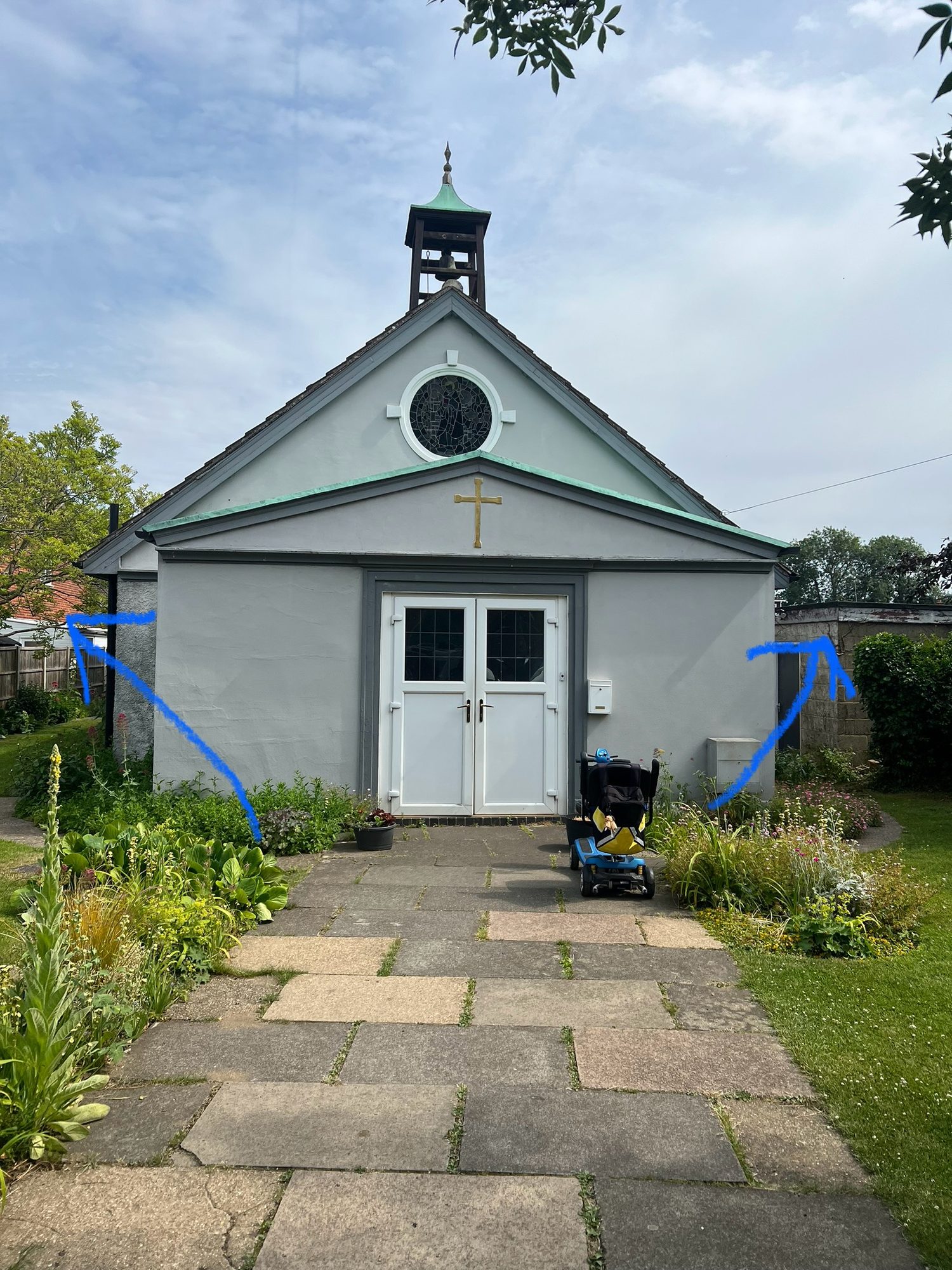 Front view of a small church with a white rendered façade, double entrance doors, a bell tower and paved pathway bordered by planted beds