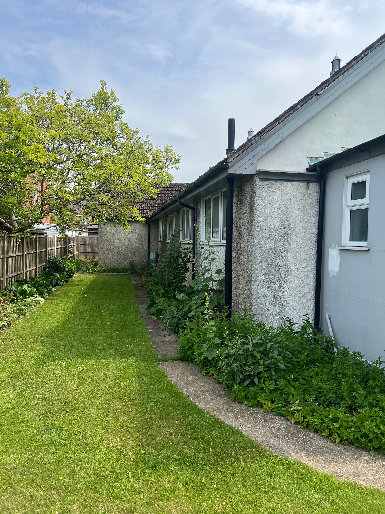 Long side view of the church showing rendered walls, multiple windows, roofline, narrow pathway and a maintained grassed area with planted borders