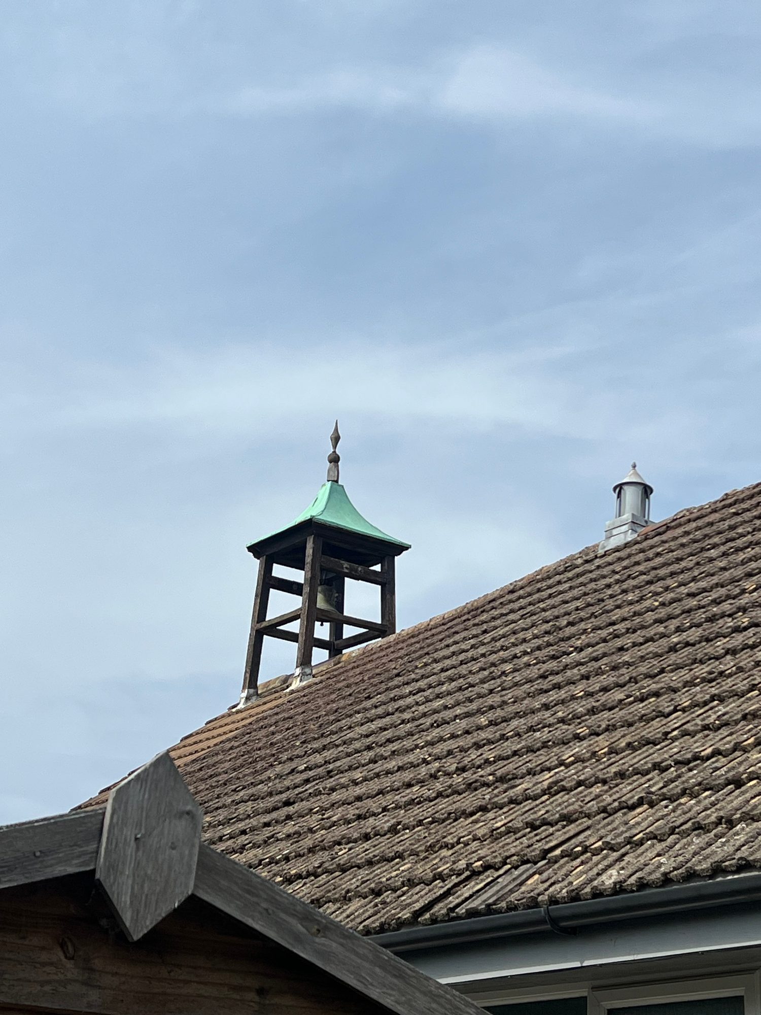 Close view of the church roof showing weathered roof tiles, guttering, a small roof vent and the timber-framed bell tower above