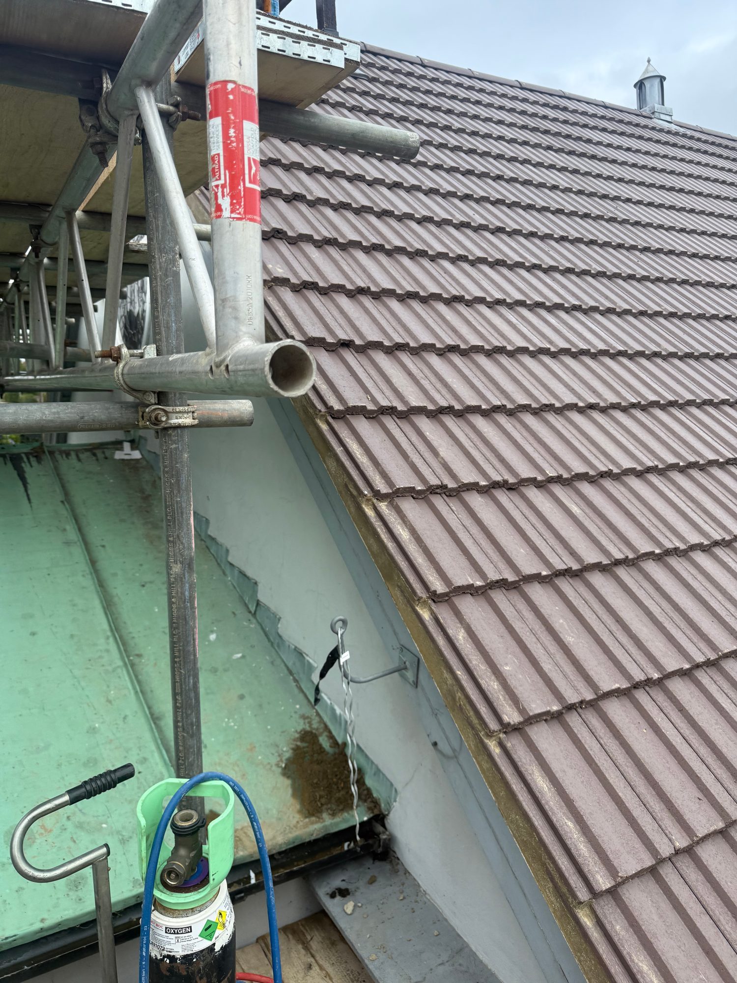 Close view of a completed pitched roof covered with evenly laid brown tiles, with roof vents visible along the slope and scaffolding beside the roof edge