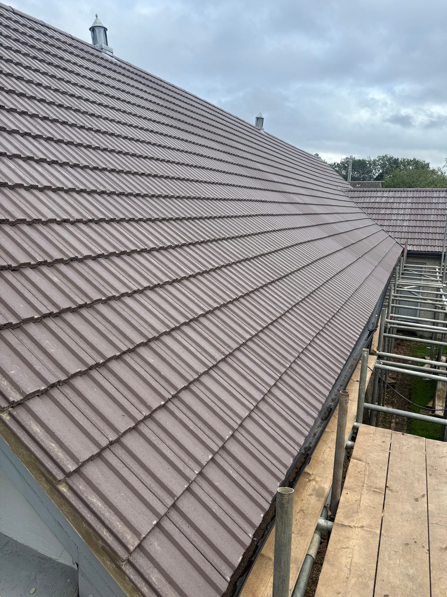 Wide view across the completed pitched roof showing rows of brown tiles installed across the main roof slope, with roof vents and scaffolding visible along the edge