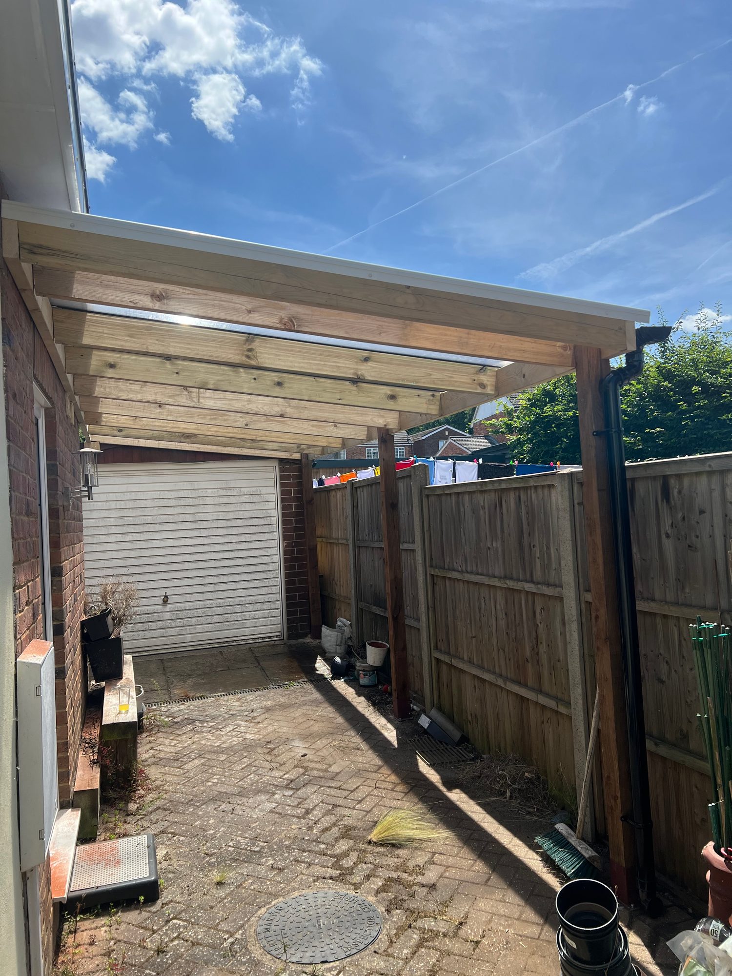 Completed garden shelter beside a brick property, showing the timber support posts, roof beams, translucent roof panels and guttering