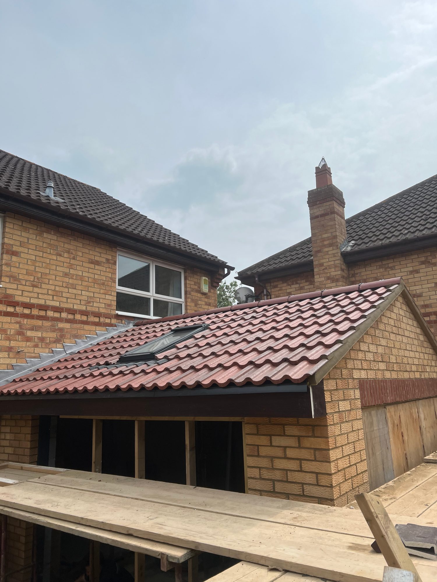 Completed red tiled garage roof with a rooflight and finished roof edges, set between existing brick buildings with scaffolding platforms visible in the foreground