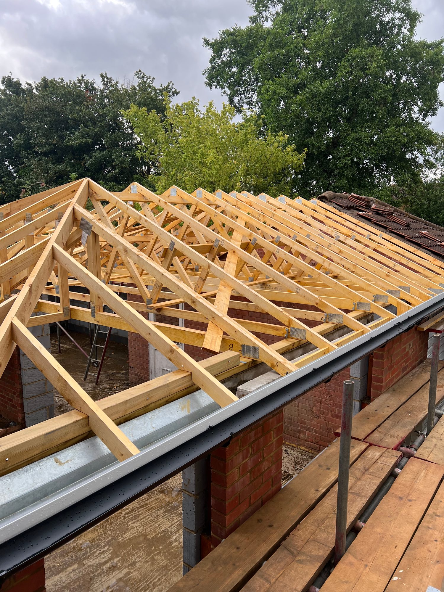Timber roof trusses installed across the building, with metal connector plates, brick and blockwork walls, scaffolding and sections of the existing tiled roof visible