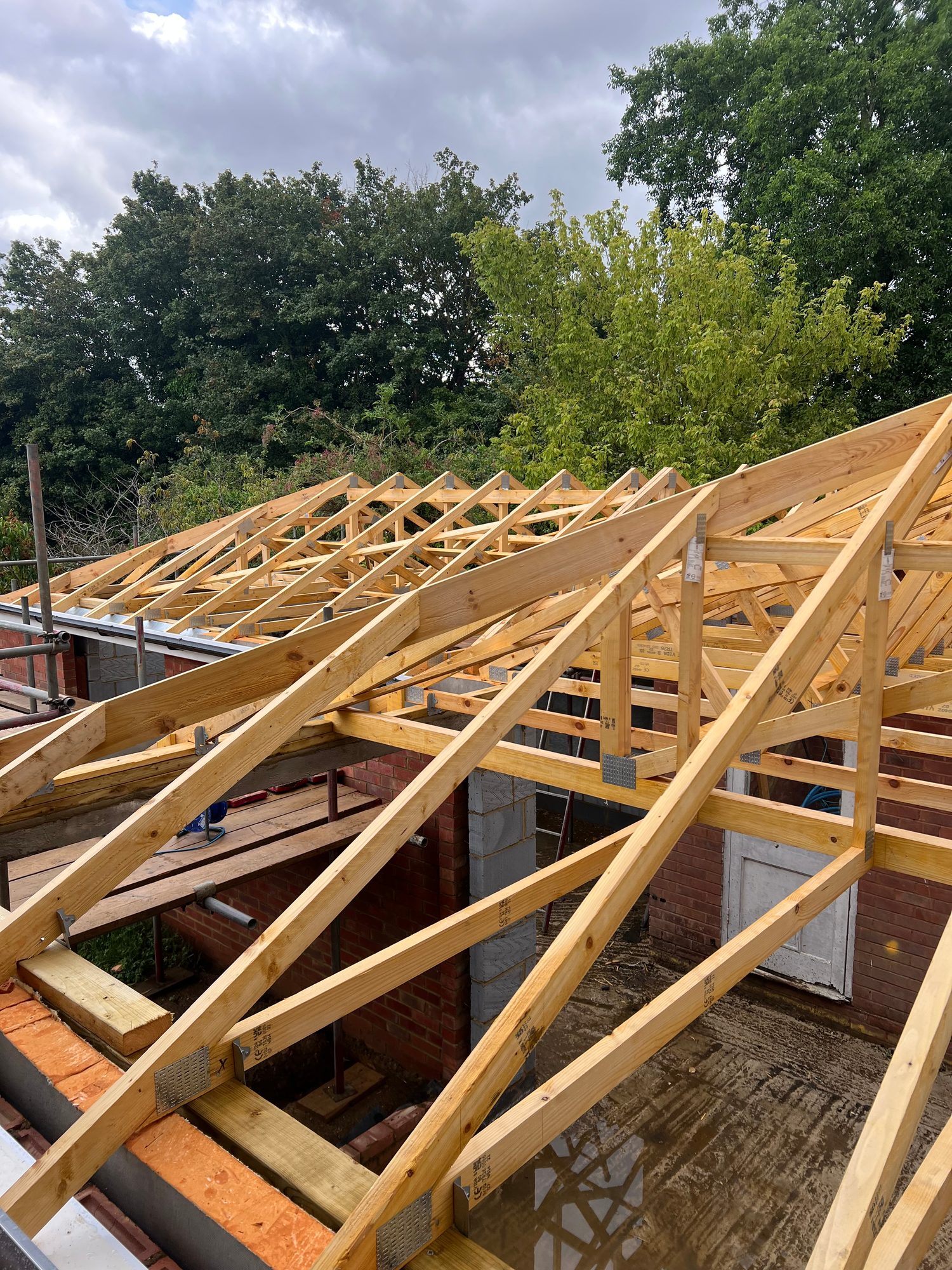 Close view of timber roof trusses and supporting roof timbers fitted across the building, with brick and blockwork walls, scaffolding and the surrounding trees visible