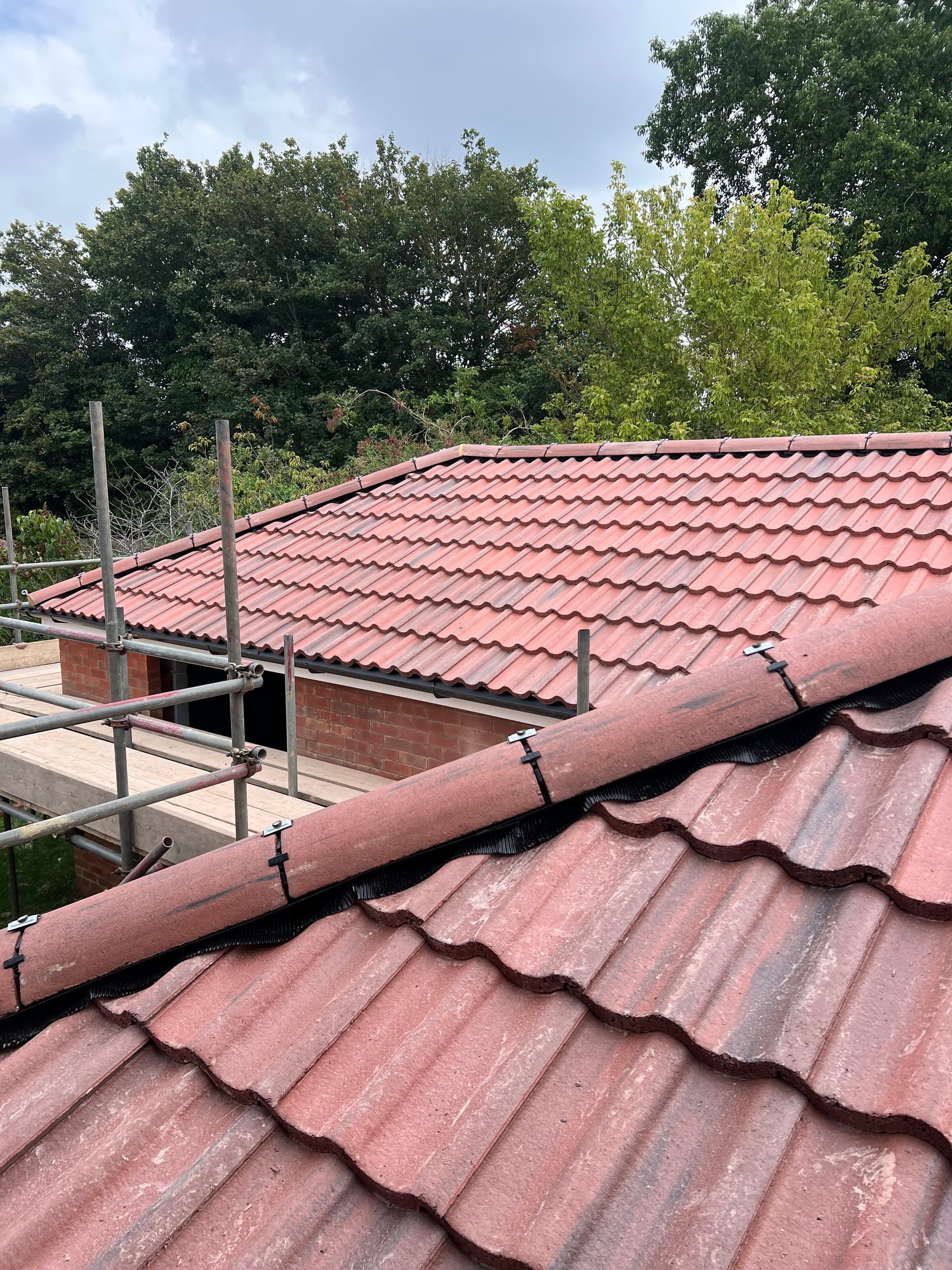 View across the completed red tiled roof showing evenly laid roof tiles and a finished ridge with black detailing, alongside the brickwork and scaffolding
