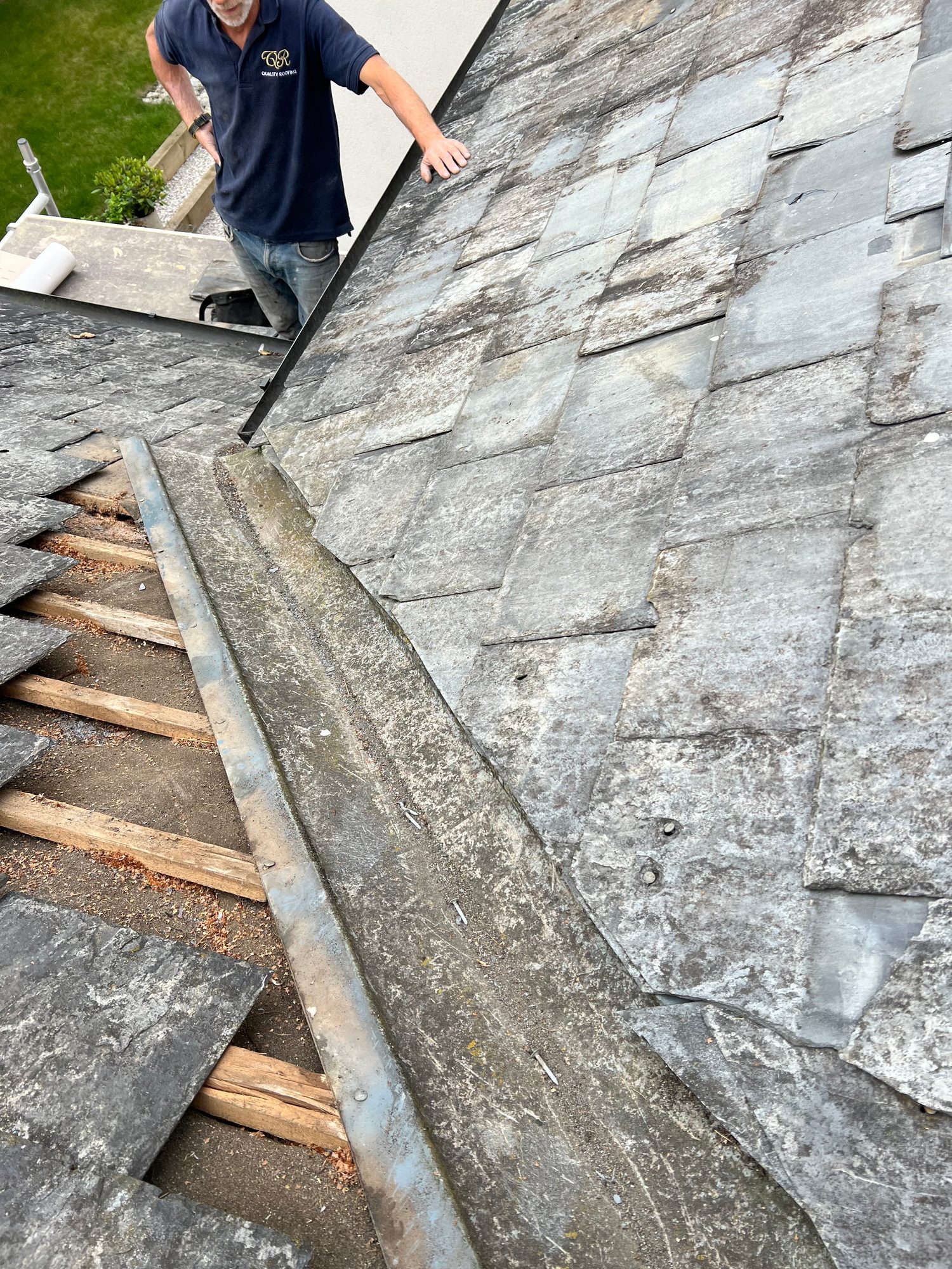 Slate roof with sections of tiles removed, exposing timber battens, while a worker is visible beside the roof