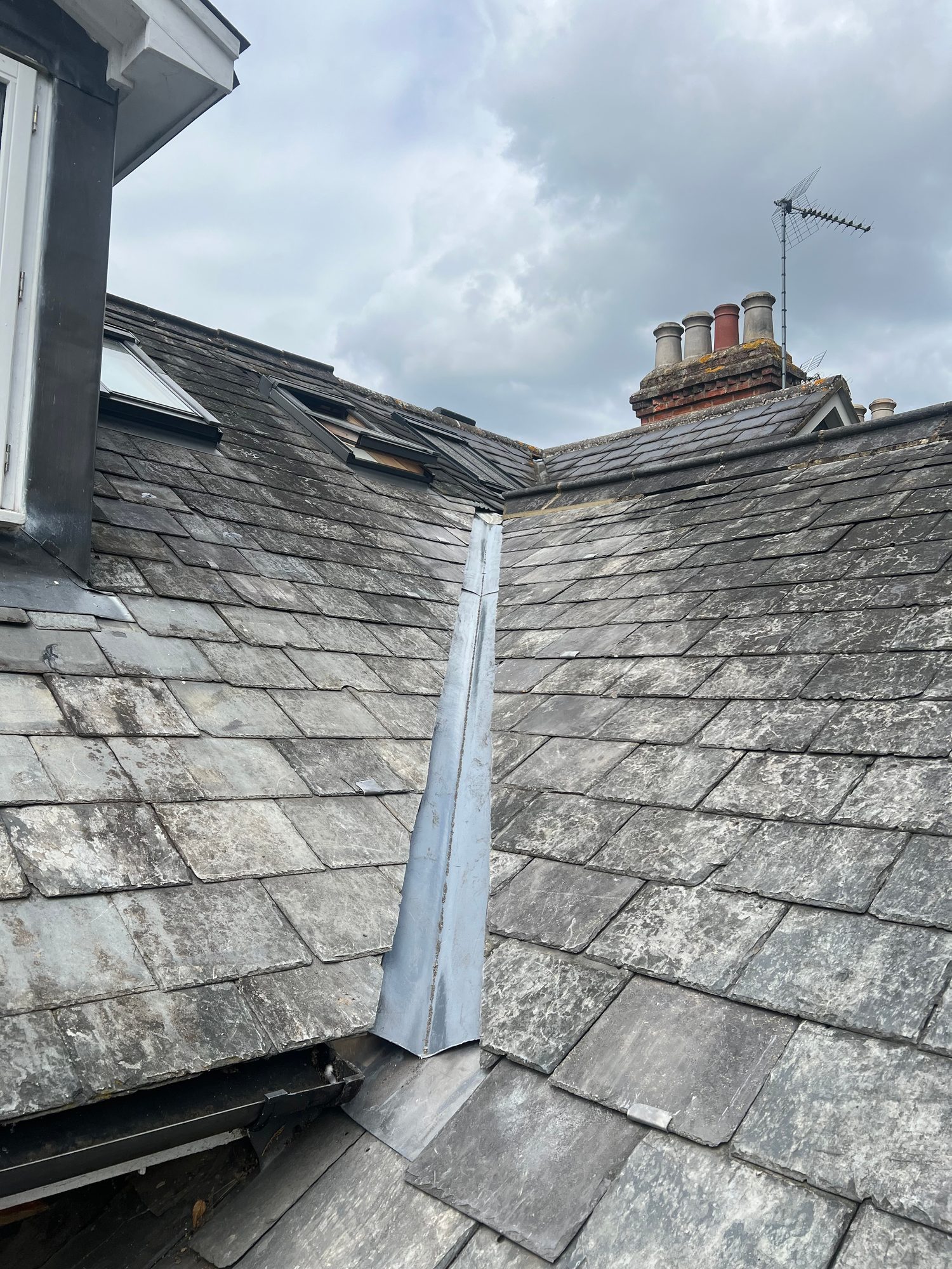 Completed slate roof showing neat rows of slate tiles and finished metal valley detailing between the roof sections, with chimney stacks visible in the background