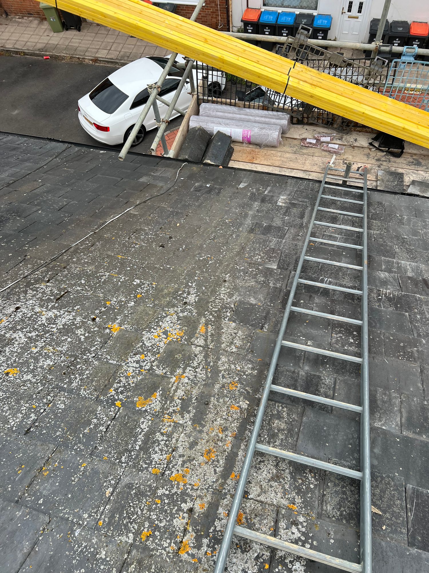 View across an existing flat roof showing weathered roof coverings, surface staining and moss or lichen growth, with access equipment and materials visible nearby