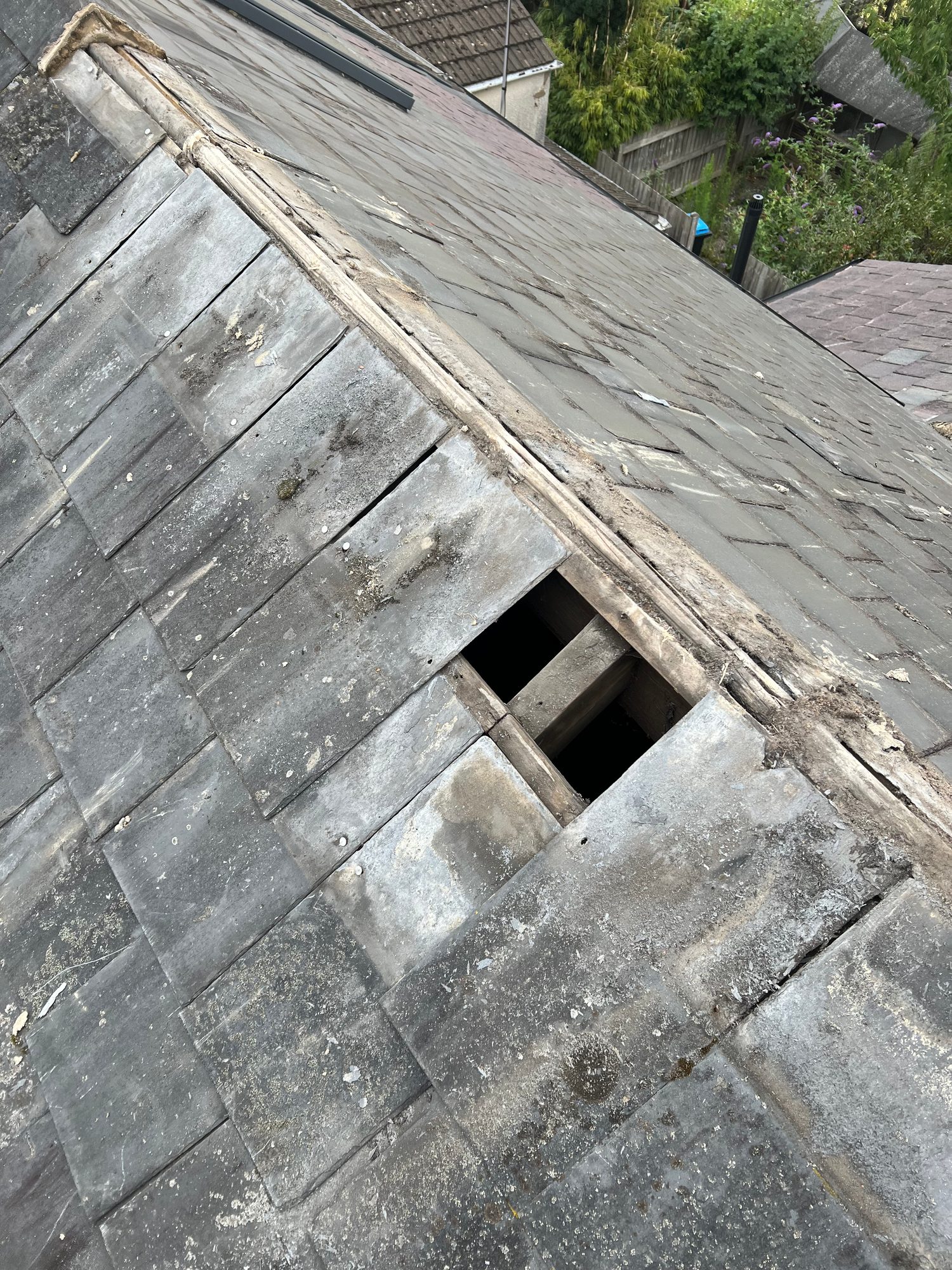 Close view of an existing pitched slate roof showing weathered and damaged slates, exposed areas beneath the roof covering and deterioration along the roof junction