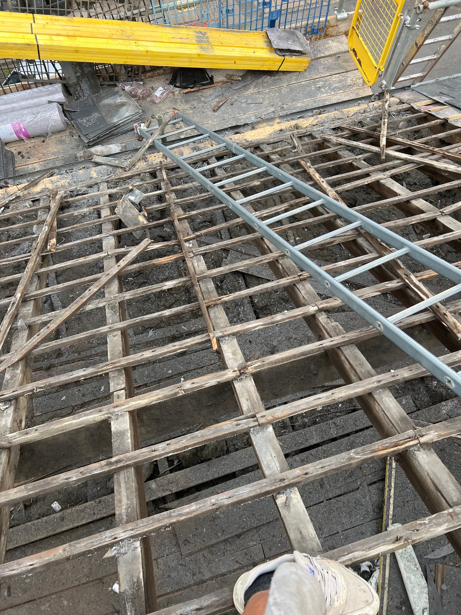 View of an exposed timber roof structure with a metal ladder laid across the timber battens and construction materials visible around the work area