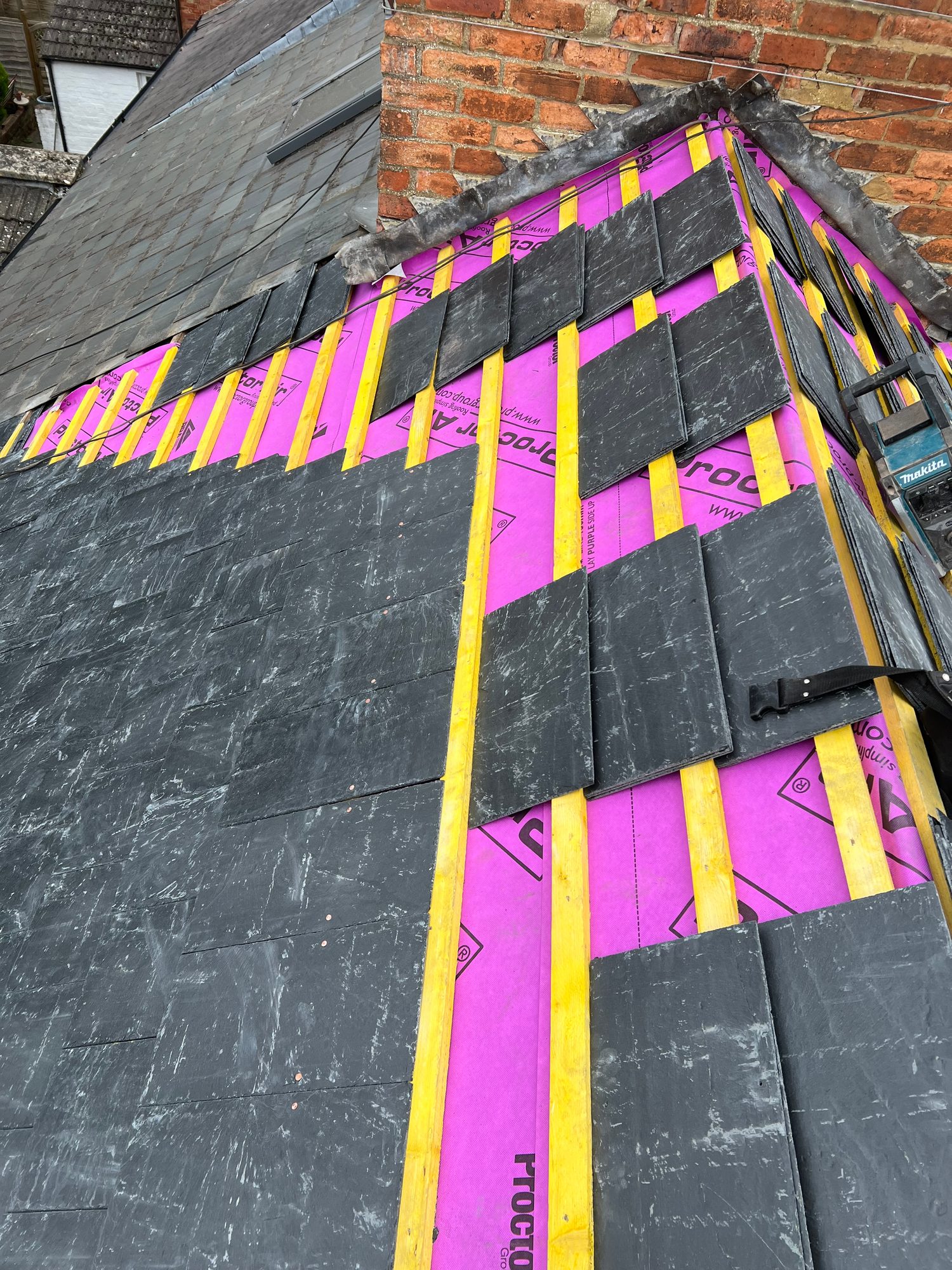 Sloping roof section showing pink insulation boards, timber battens and partially installed slate tiles alongside a brick wall