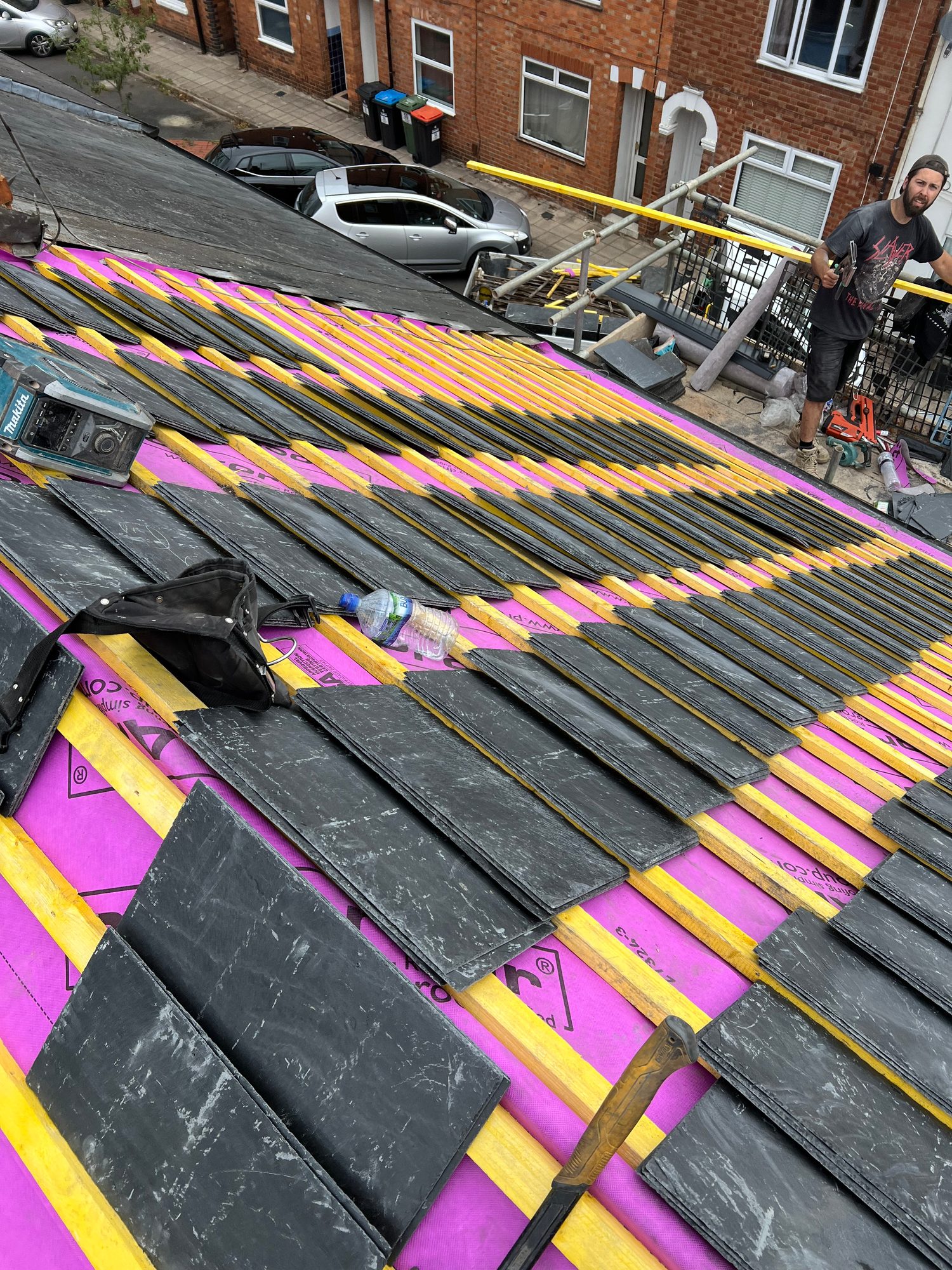 Rows of dark slate tiles laid across timber battens and pink roofing underlay, with roofing tools and scaffolding visible around the work area