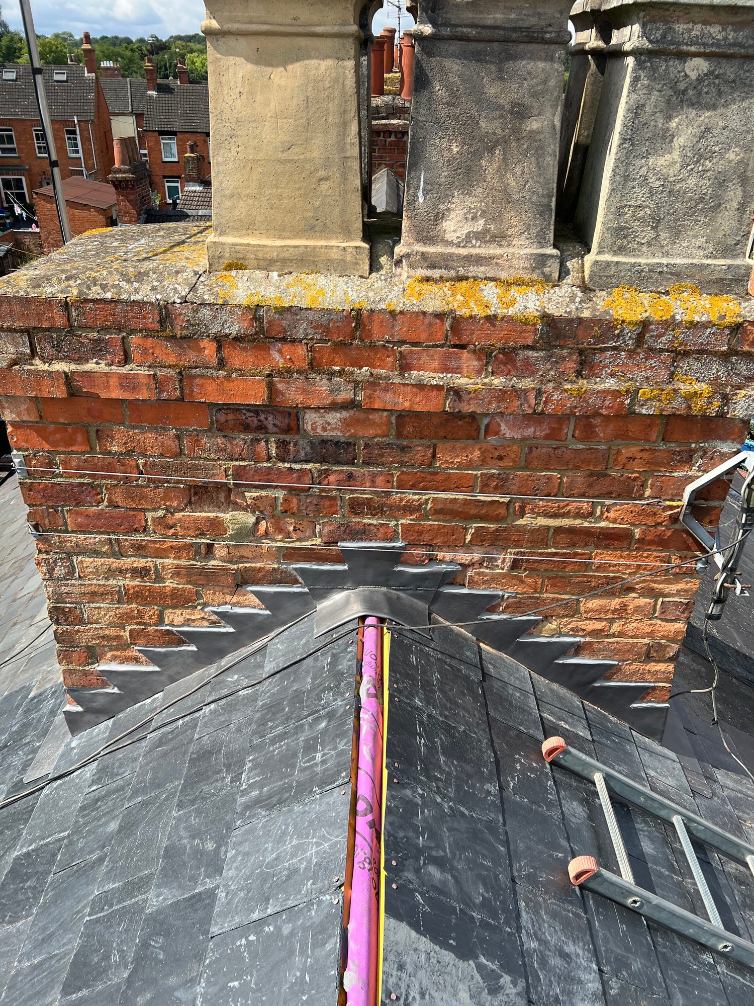 Dark slate roof covering meeting a brick chimney, with stepped lead flashing visible around the chimney base and roof junction