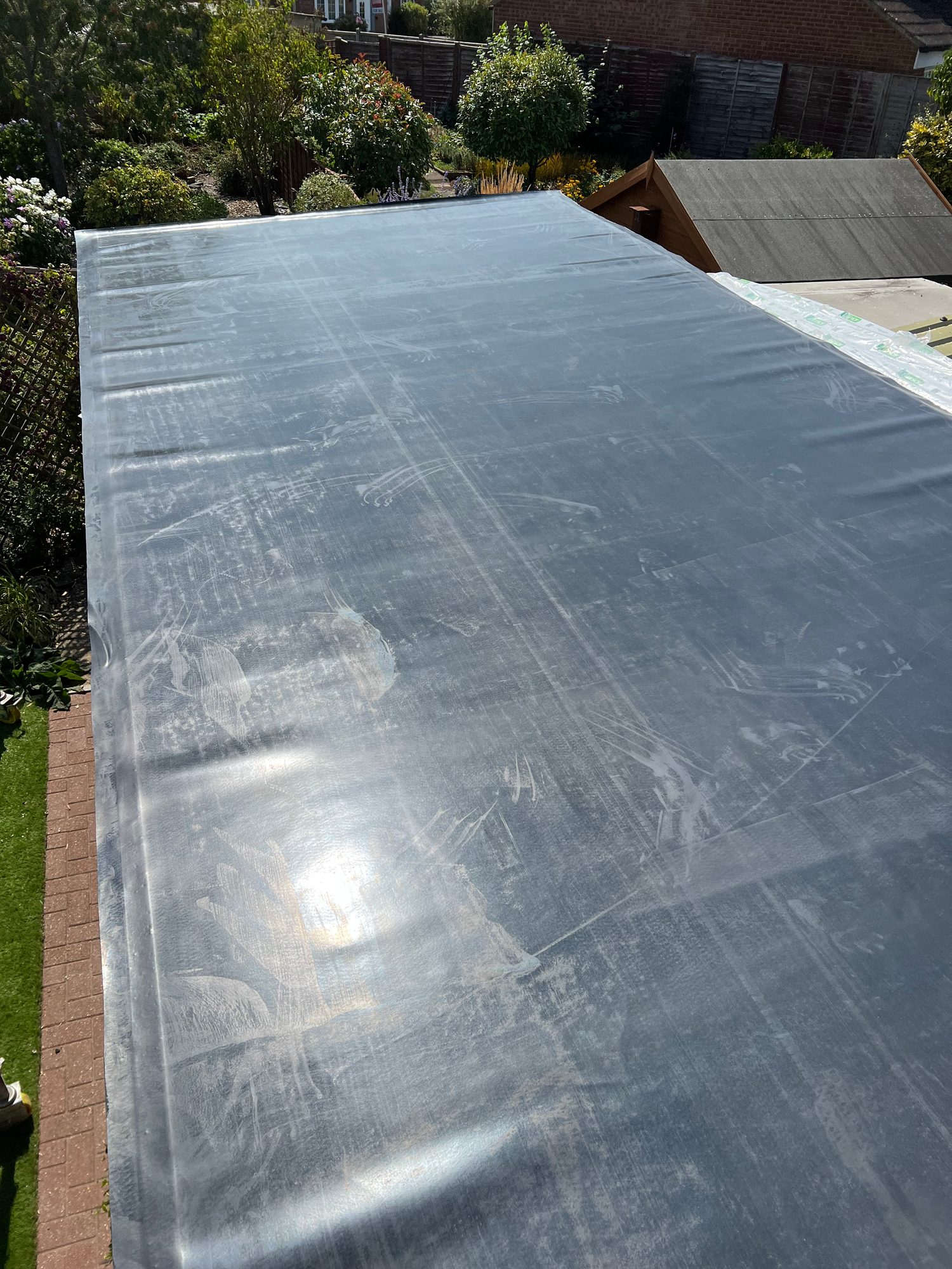 Close-up view of a roof covering with visible surface marks and reflections, with a garden and adjacent outbuilding in the background