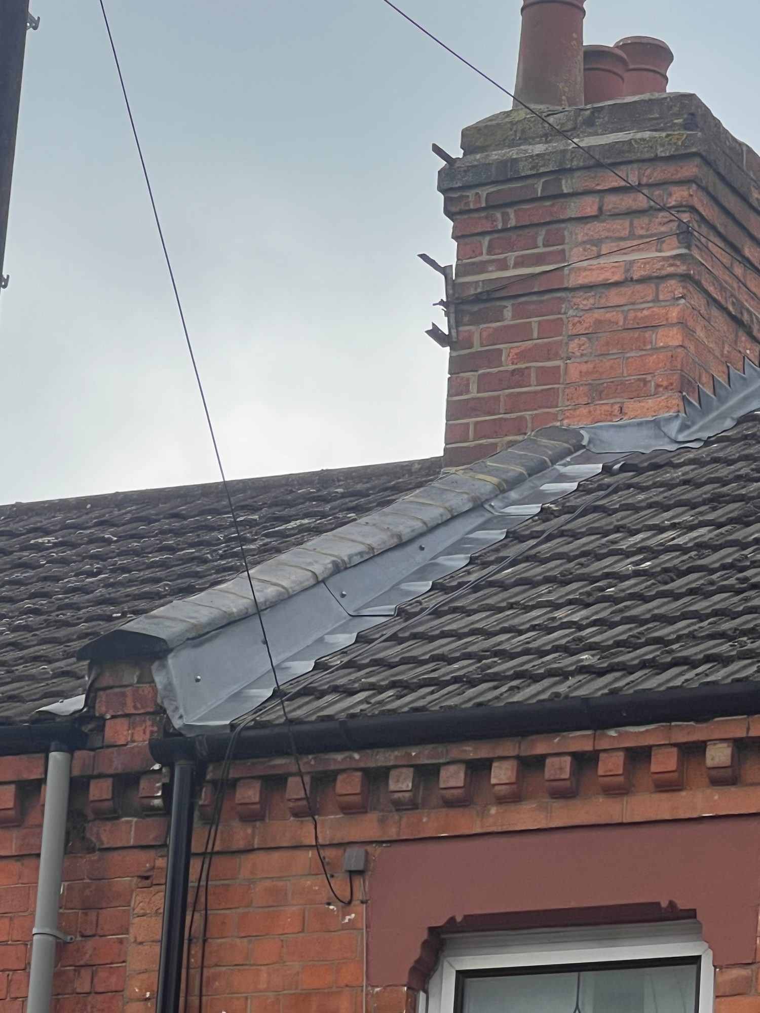 ComRoof valley with new metal flashing installed between tiled roof slopes and around the brick chimney, with the gutter and downpipe visible belowpleted