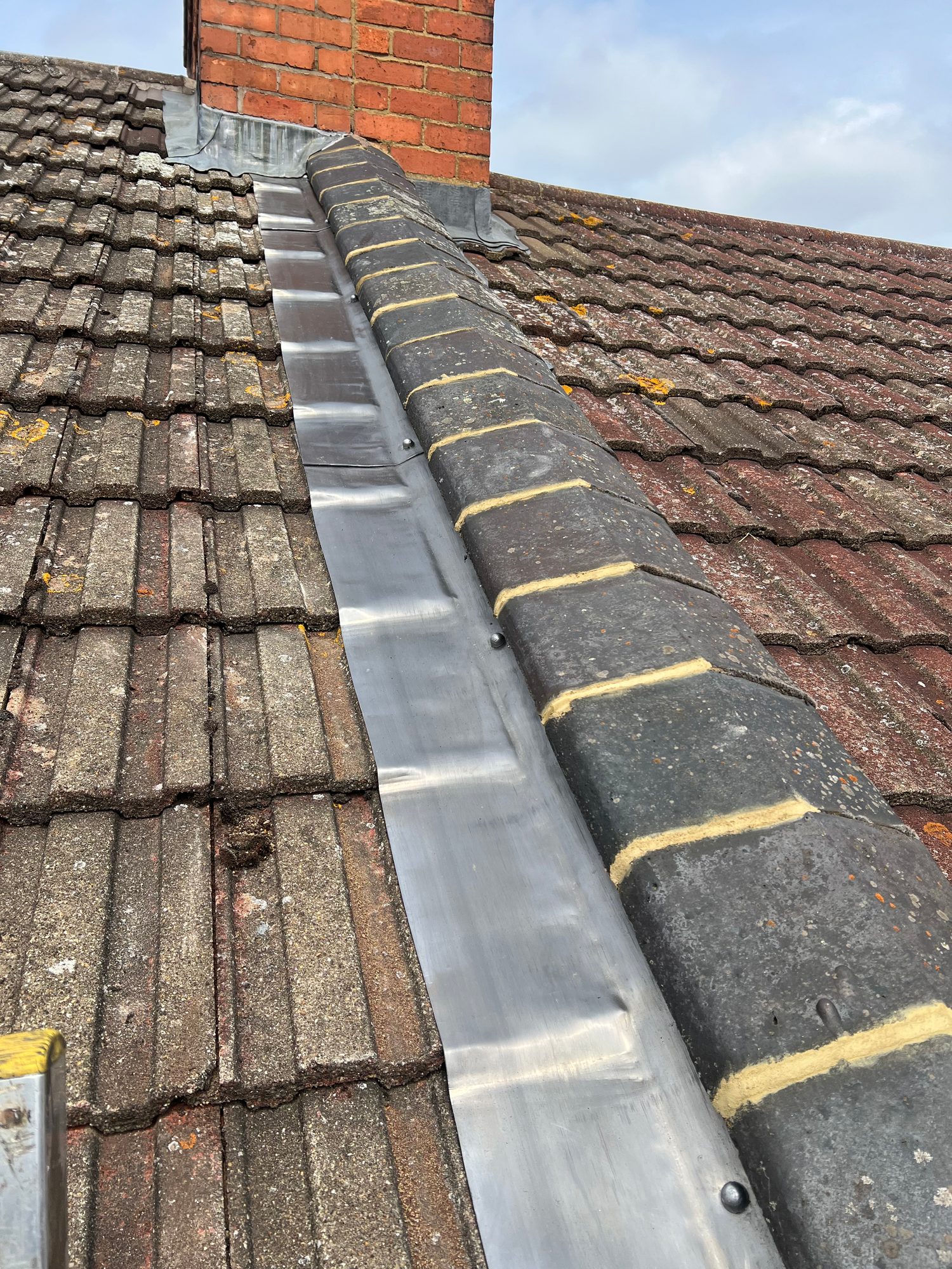 Close-up of metal flashing installed along the roof valley beside the tiled roof slopes and beneath the brick chimney, with fixings visible along the flashing