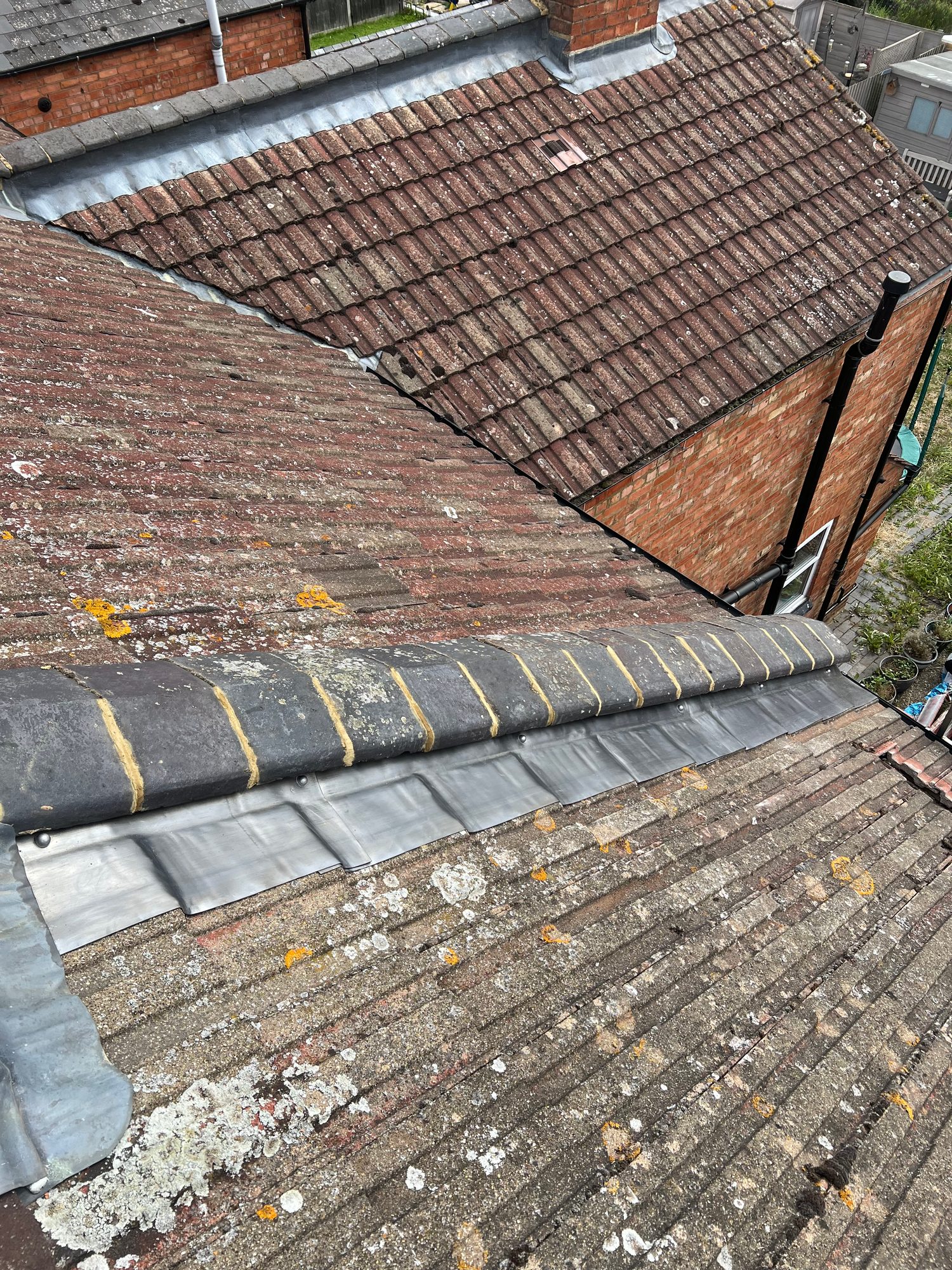 Wide view of a tiled roof showing newly installed metal flashing along the roof valley and beneath the ridge tiles, with the brick chimney and roof drainage visible