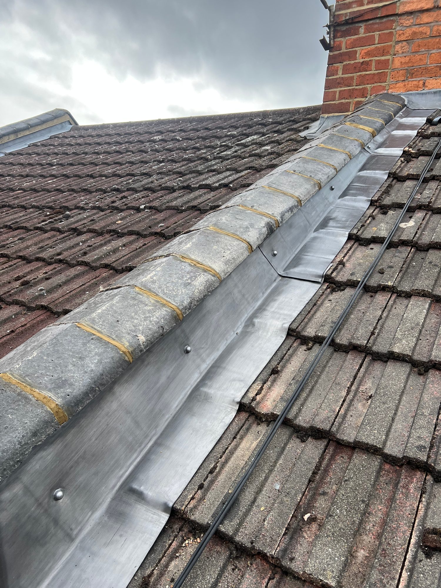 Close-up view of metal flashing securely fixed along the roof valley beside the tiled slopes and extending up to the brick chimney