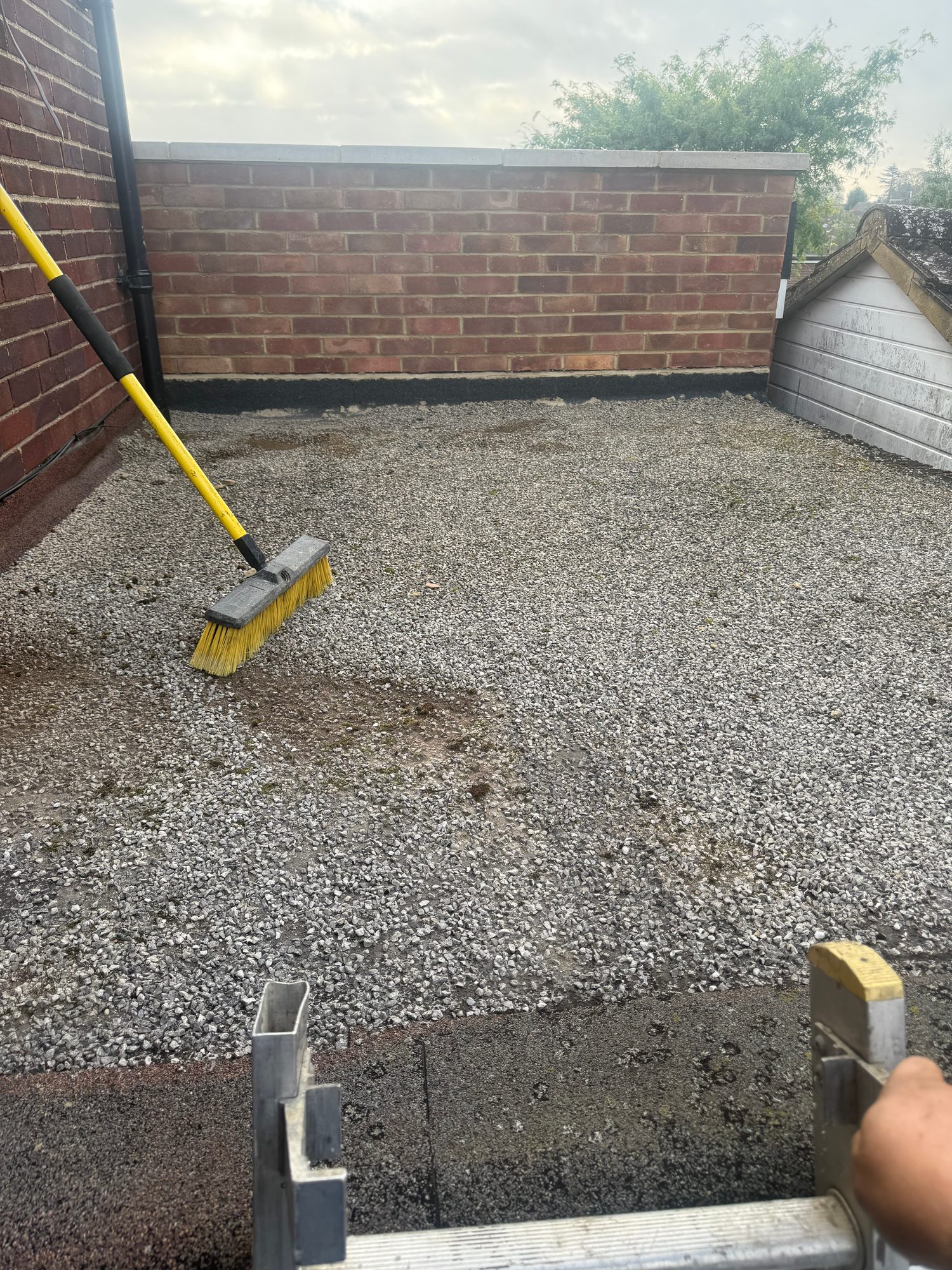 Flat roof covered with loose stone aggregate, with a roof brush and access ladder visible in the foreground and brick parapet walls around the roof perimeter
