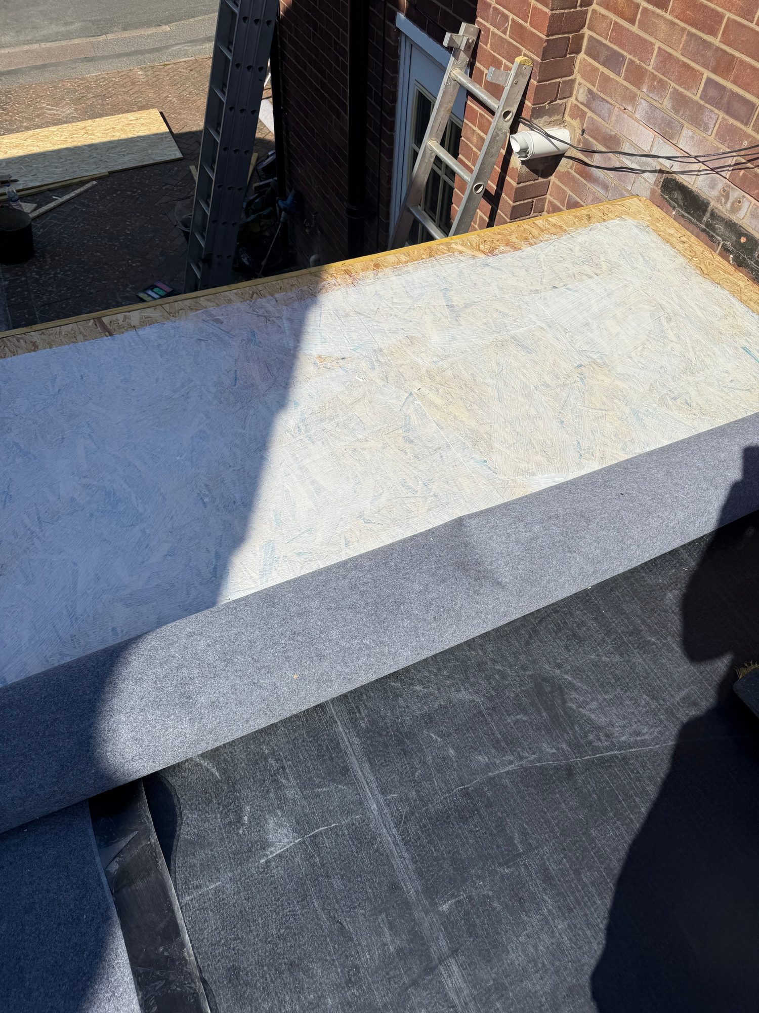 Close-up of a flat roof during refurbishment, showing a dark roof surface, grey membrane material and exposed board beneath, with ladders and roof access equipment visible nearby