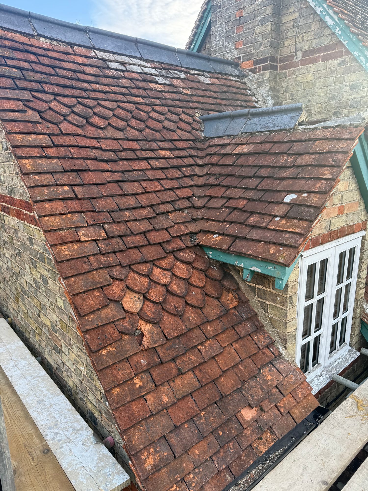 Close-up view of the existing tiled roof showing weathered tiles, decorative tile sections, roof junctions and exposed roof edging around the brick building
