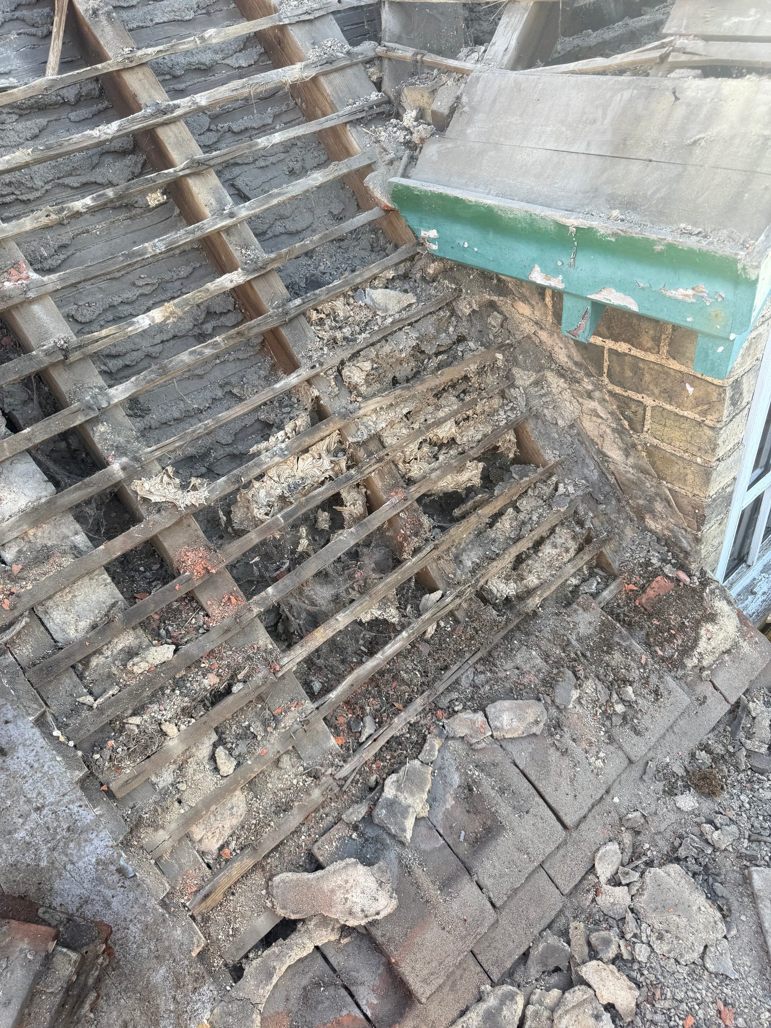 Roof area during construction with tiles removed, exposing timber battens, roof structure and underlying masonry, with debris and remaining roof tiles visible around the work area