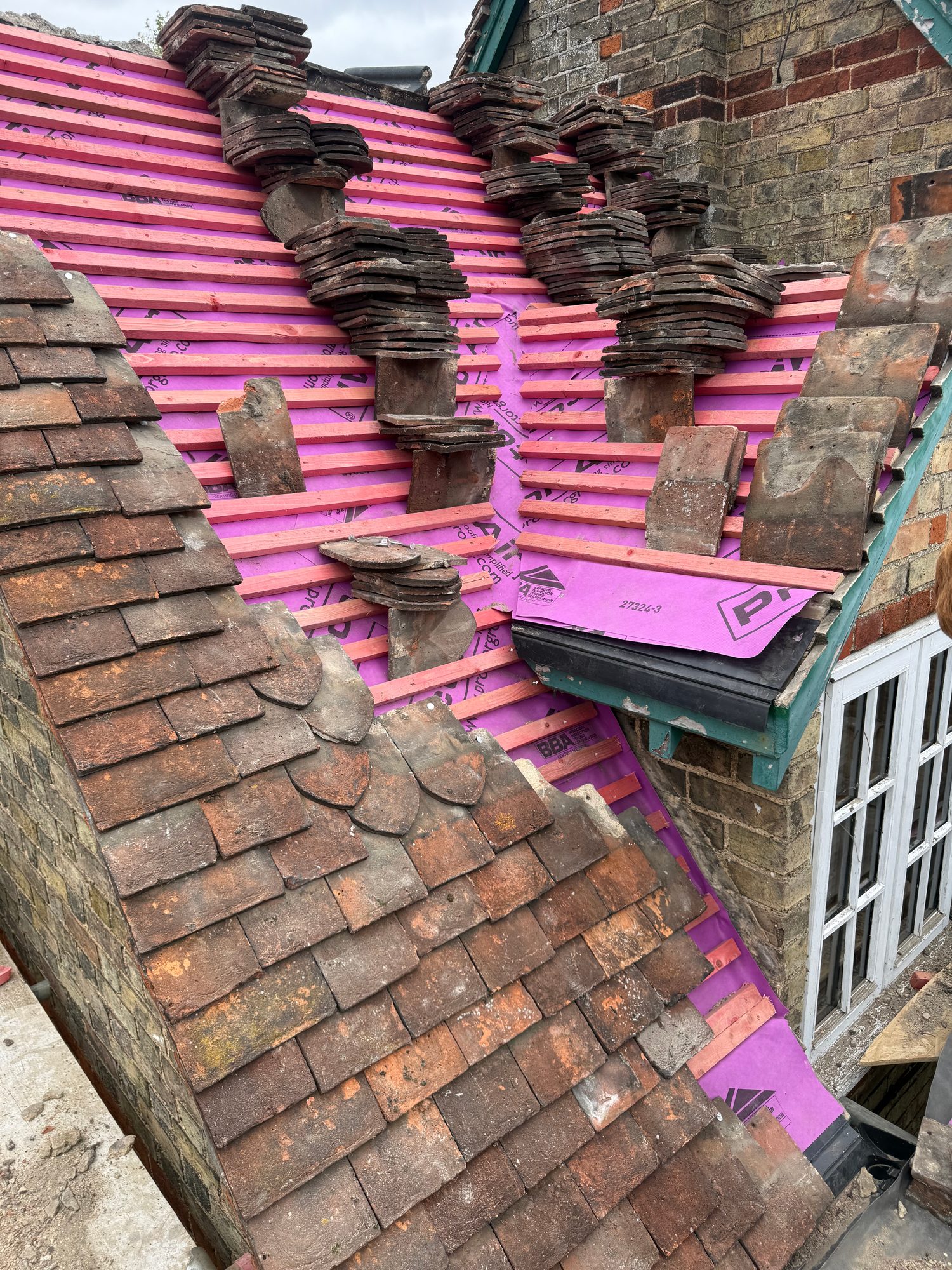 Roof during construction showing pink insulation membrane and new timber battens installed around existing chimney stacks, with roof tiles stacked nearby and sections of the original tiled roof remain