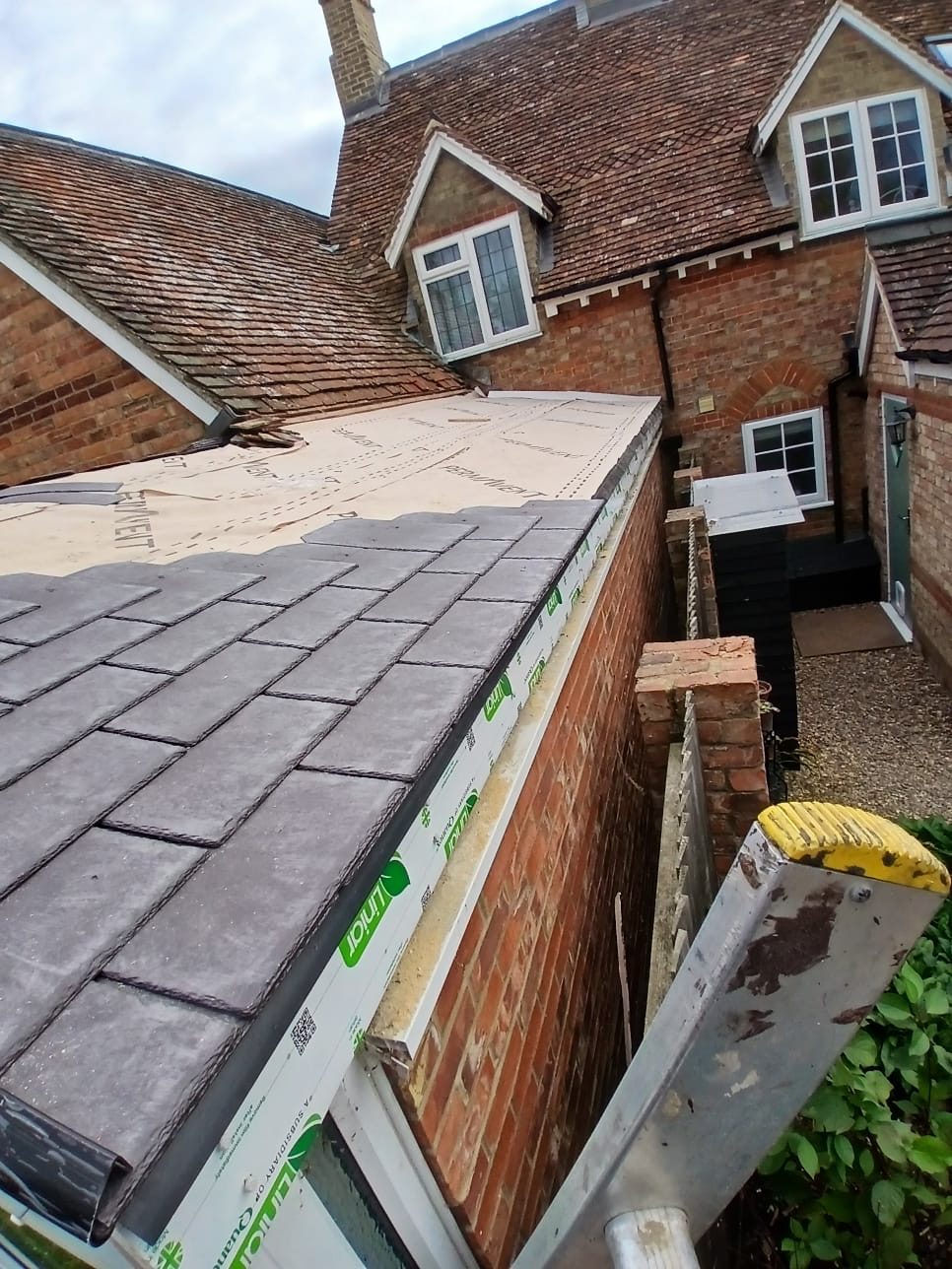 Partially completed roof with new roof tiles and exposed roofing materials along the edge, with an access ladder beside the brick structure