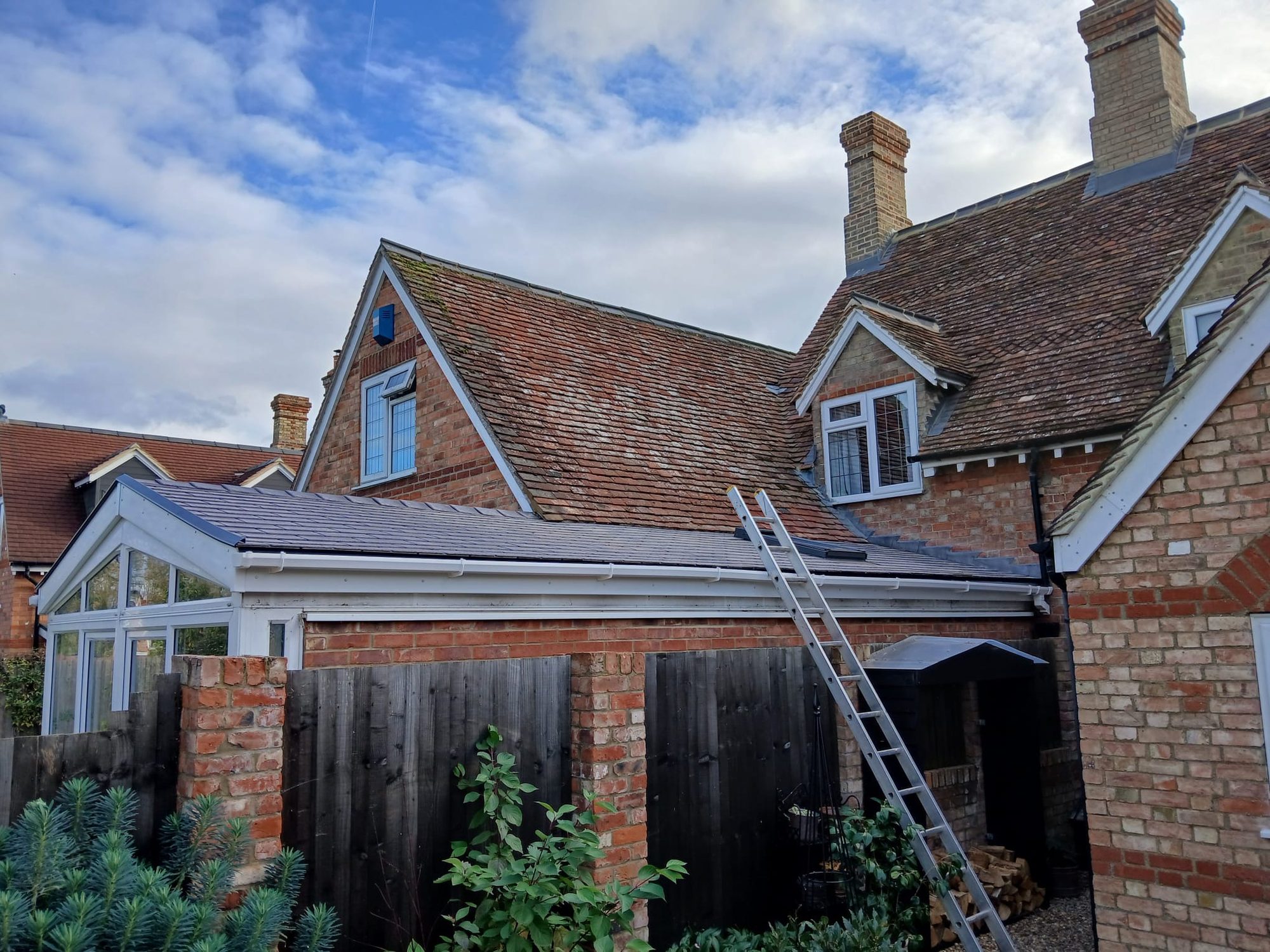 Completed tiled roof with white roof edging and guttering above the brick conservatory, with an access ladder positioned against the roof and surrounding brickwork visible