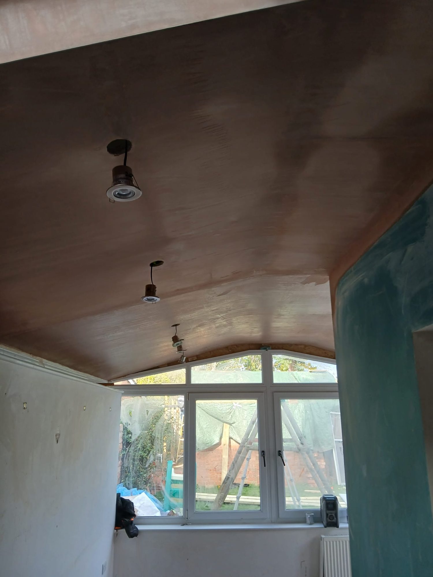 Interior view during construction showing a partially finished curved ceiling with recessed light fittings being installed, white-framed windows and unfinished wall surfaces visible below