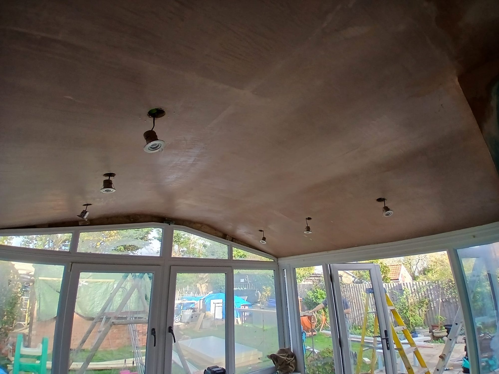 Interior view showing a curved ceiling with multiple recessed light fittings in place, alongside white-framed windows and doors opening onto the garden
