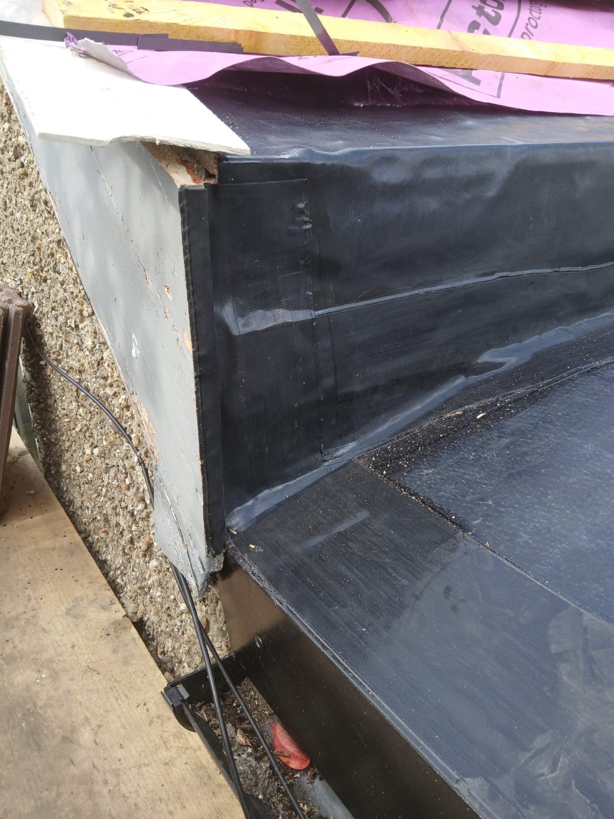 Close-up of the flat roof edge showing black waterproofing material, flashing and purple roofing underlay at the junction with the roof structure